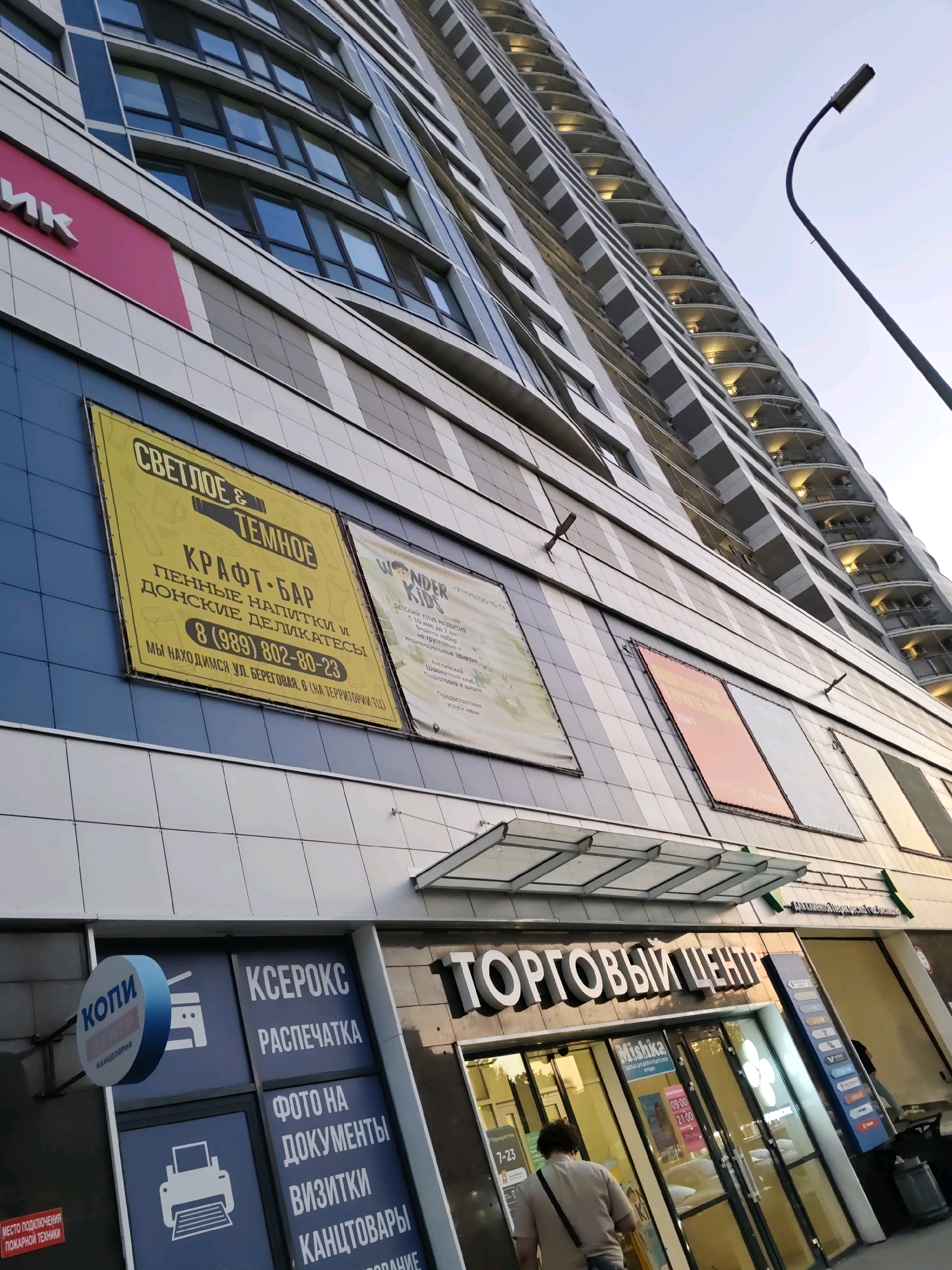 The image shows a modern, multi-story building in Rostov-on-Don, Russia. The building features a facade of white and grey tiles with blue accents, and numerous windows with reflective glass. On the lower levels, there are several commercial establishments. Prominently displayed are large yellow and white signs for "Светлое & Темное" (Craft Beer & Bar) and "Wonder Kids". Adjacent to these, a shop named "Копи- Центр" (Copy Center) has a circular blue and red sign. Further down, the entrance to a "Торговый Центр" (Shopping Center) is visible, with glass doors and display windows. A person is seen walking towards the shopping center entrance. The overall environment suggests an urban commercial district.