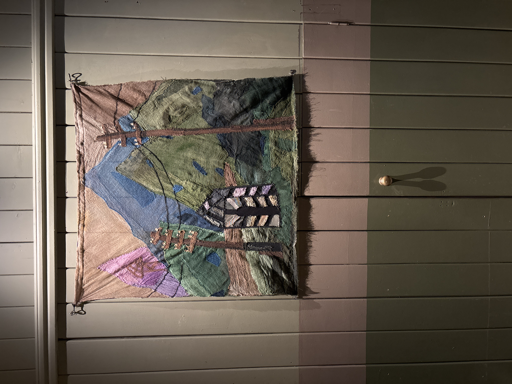 A square-shaped fabric art piece, resembling a quilt or tapestry, hangs against a wooden paneled wall. The artwork depicts a stylized landscape with abstract forms in various shades of green, blue, brown, and hints of purple and pink fabric patches. Notable elements within the art include a winding blue section, possibly a river, and horizontal brown bands that traverse the scene. The upper right section of the artwork features textured black and lighter-toned rectangular fabric blocks.
The background wall is composed of horizontal wooden panels, painted in two distinct vertical sections: a lighter grey-green on the left, encompassing the hanging art, and a darker green on the right. The art piece is suspended by four metal hooks, two of which are visible in the upper corners. A brass-colored door handle or knob is located on the darker, right-hand side of the wall, casting a prominent, elongated shadow to its right.
The scene is illuminated by a focused light source from the left, brightly highlighting the fabric art and the lighter section of the wall, while casting the right side of the wall and the door handle into deeper shadow. The distinct horizontal paneling of the wall suggests a traditional architectural style. No visible text is present in the image.