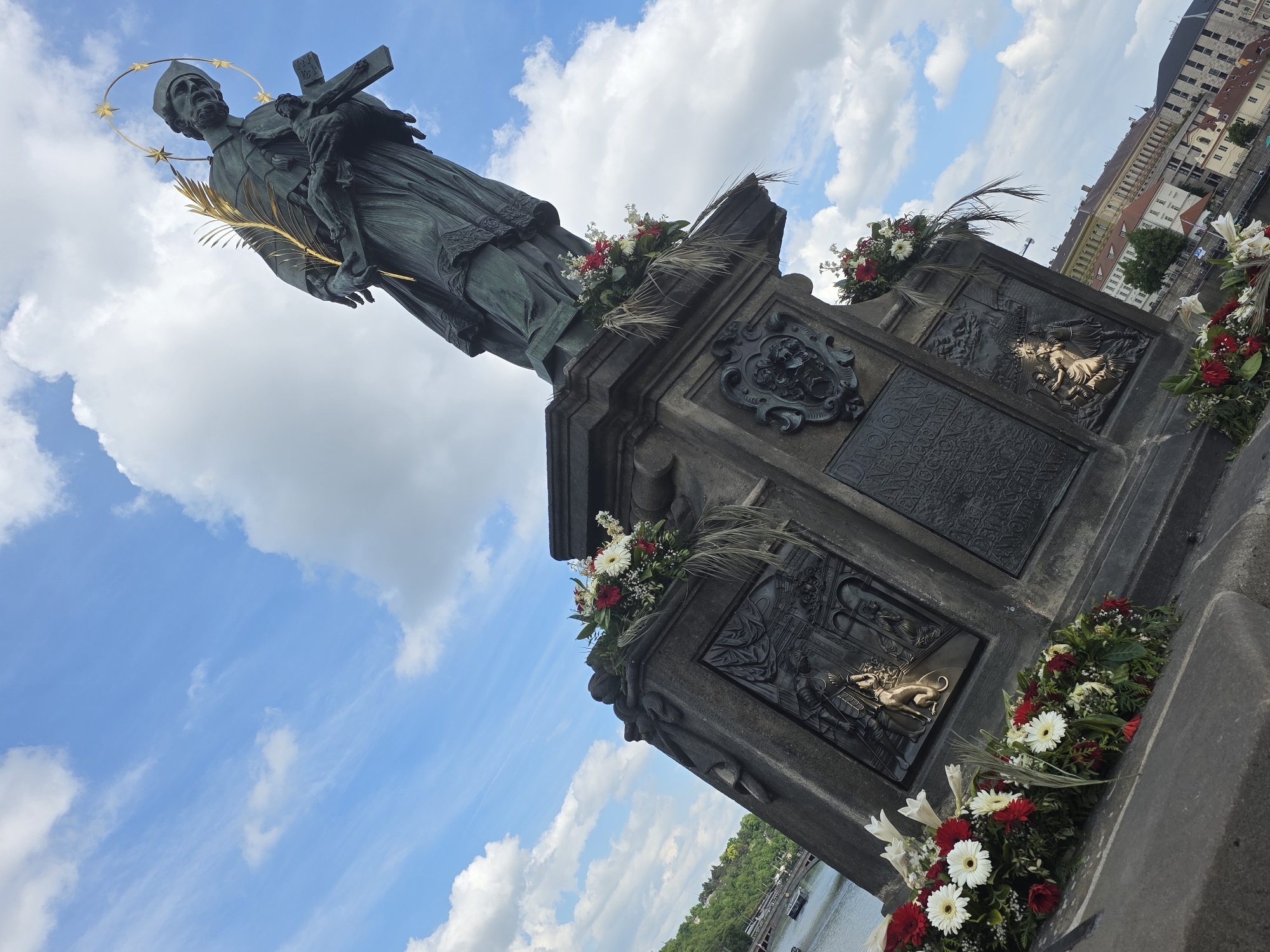 The image depicts a bronze statue of a saint holding a cross with Jesus crucified. The statue stands atop a gray stone pedestal decorated with flowers and bronze plaques. The sky in the background is blue with white clouds. The city of Prague, Czech Republic can be seen in the background.