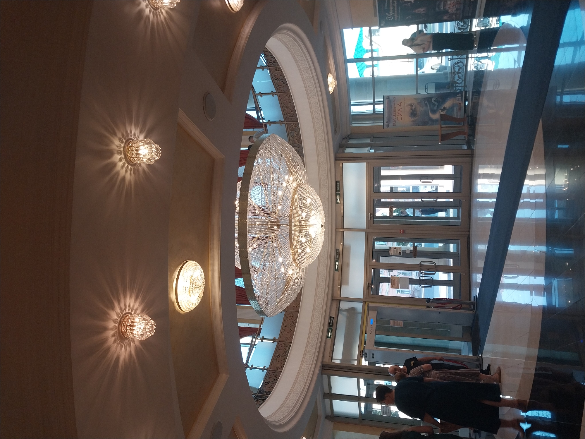 The image depicts the interior of a building with grand architecture, likely a public space such as a theater or a shopping mall. A large, ornate crystal chandelier hangs prominently in the center of the frame, casting a warm glow. Several smaller sconce lights with intricate glass details are visible on the walls, illuminating the space with radiating patterns of light. The scene is well-lit, suggesting it's daytime, though the interior lighting plays a significant role.
The architecture features elegant arches and decorative moldings, contributing to a luxurious ambiance. Large glass doors are visible in the background, with reflections on the polished floor creating a sense of depth. Through the doors, one can glimpse an outdoor area with buildings and windows, hinting at an urban setting.
Several people are present in the image, though their faces are not clearly visible due to the image's orientation and focus. They appear to be walking or standing, engaged in normal activity. One individual is seen near a metal detector frame, suggesting a security checkpoint. A poster with Cyrillic text is displayed near the entrance. The text on the poster reads "ВОЛШЕБНАЯ СИЛА" which translates to "Magical Power." Another visible sign near the doors reads "ВЫХОД" meaning "Exit." The overall impression is one of a sophisticated and busy indoor environment.