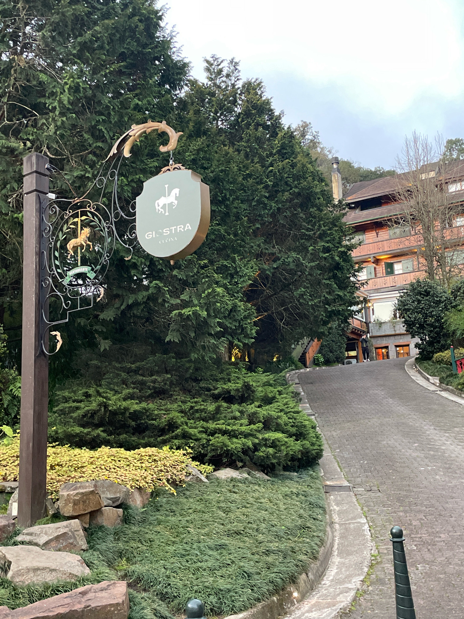 The image depicts the entrance to "Gioastra Cucina" restaurant, located in the city of Gramado, Brazil. The scene is set outdoors with a paved driveway leading towards a multi-story building with wooden facade and balconies, suggesting an alpine-style architecture characteristic of Gramado. On the left side of the driveway, a decorative signpost features an ornate metal bracket supporting two signs. The upper sign is shield-shaped and green, bearing the text "GIOASTRA CUCINA" and an image of a white horse. Below this, a circular sign with a golden horse figure is visible. The surrounding environment consists of lush green trees, manicured bushes, and large rocks, creating a natural and somewhat rustic atmosphere. The sky is overcast, indicating a possibly cool or temperate climate. A person in dark clothing is partially visible walking up the driveway towards the building.