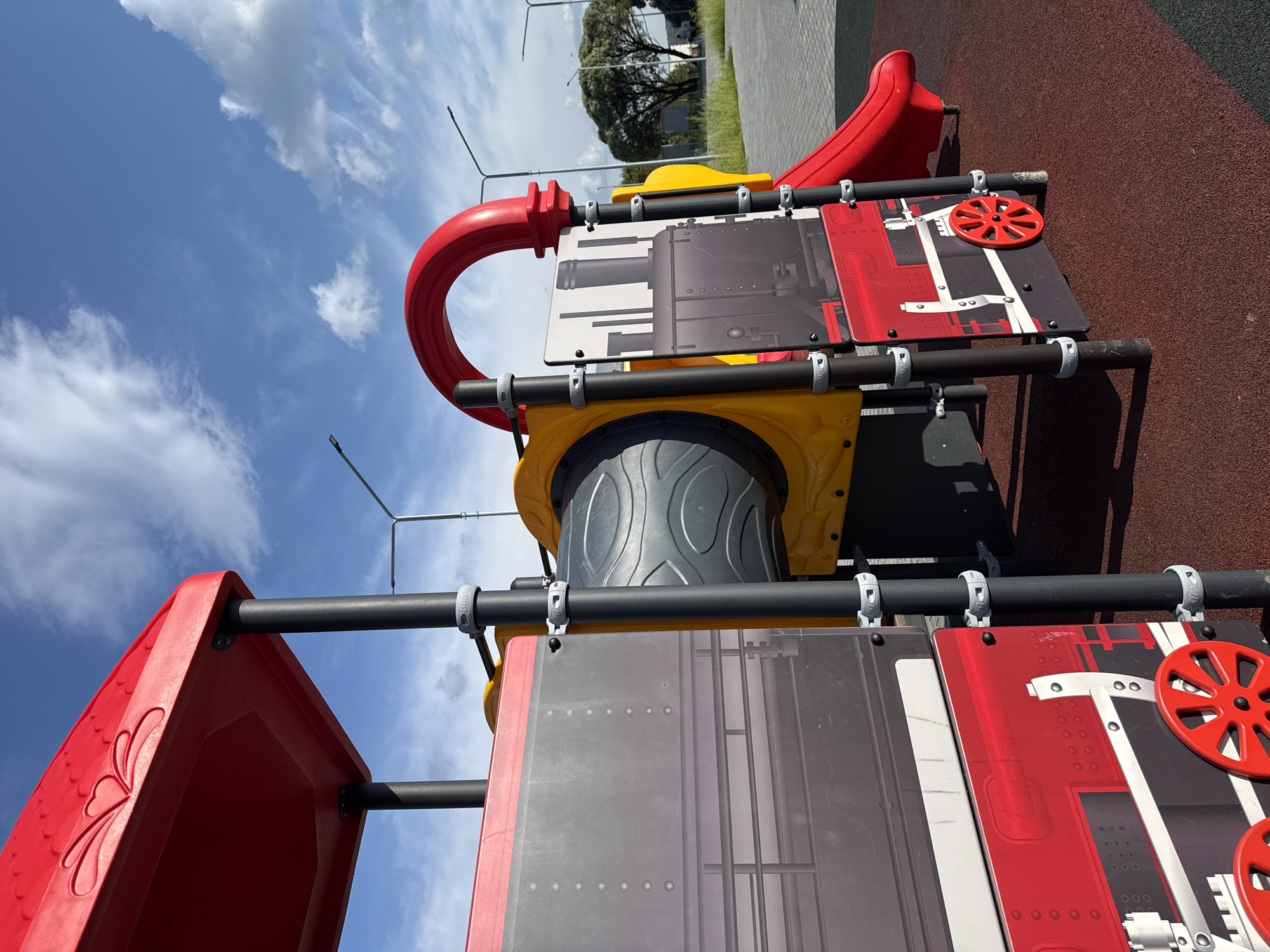 A close-up, low-angle shot captures a section of a red and yellow children's playground structure designed to resemble a train, set against a bright blue sky with scattered white clouds. The playground equipment features black support poles, red decorative elements with heart shapes on the upper structure, and a red slide. One side of the train structure displays graphics of train windows and wheels, with gray panels depicting a train engine. The ground beneath the playground is a textured, dark red rubberized surface. In the background, a paved area, streetlights, a few trees, and a hint of grass are visible, suggesting an outdoor urban setting. This scene is located in Vereshchagino, Russia. No people are present in the immediate vicinity of the playground.