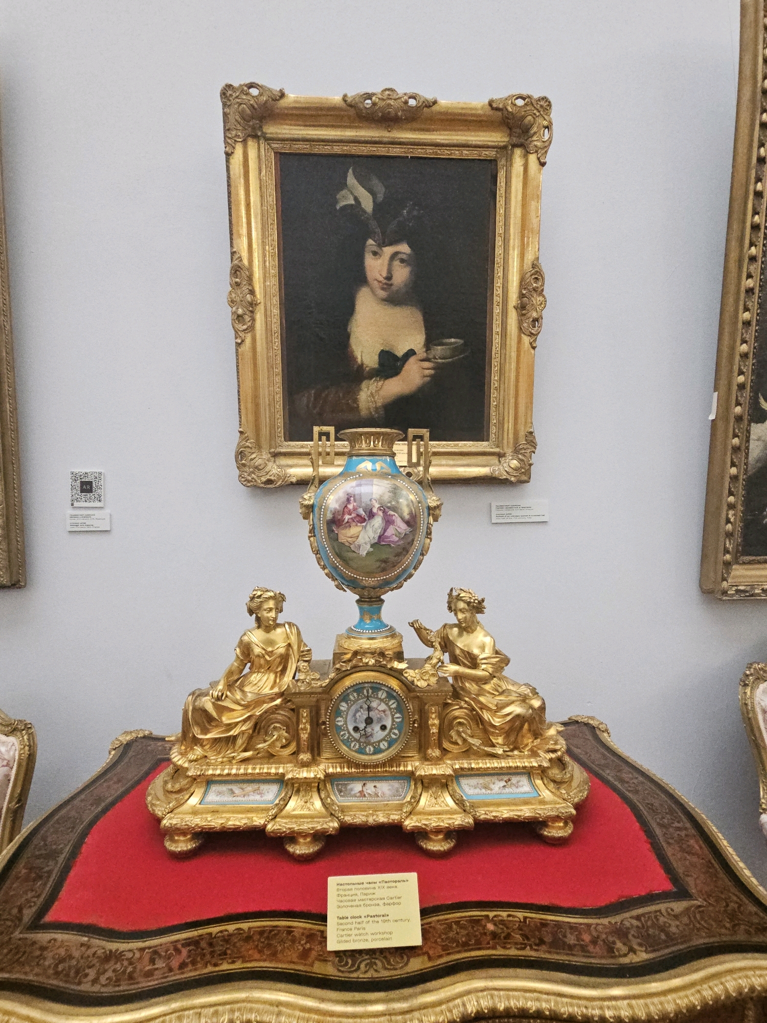 The image shows an ornate gilded bronze and porcelain table clock in the foreground, placed on a red velvet draped surface. The clock features two seated female figures, one on each side of the clock face, which is decorated with blue and white porcelain elements. Above the clock is a decorative porcelain vase with painted scenes. Behind the clock and vase hangs a framed oil painting of a woman holding a cup. To the left of the painting, a QR code and a label are attached to the wall. To the right of the central painting, part of another framed painting is visible. The environment appears to be a museum setting, with light gray walls and other exhibition elements visible on the sides and bottom of the frame. The label on the clock reads "Table clock «Pastoral» Second half of the 19th century. France Paris. Cartier watch workshop. Gilded bronze, porcelain". Based on the provided information and the presence of the label, this artifact is displayed in the Ulyanovsk Regional Art Museum, located in Ulyanovsk, Russia.