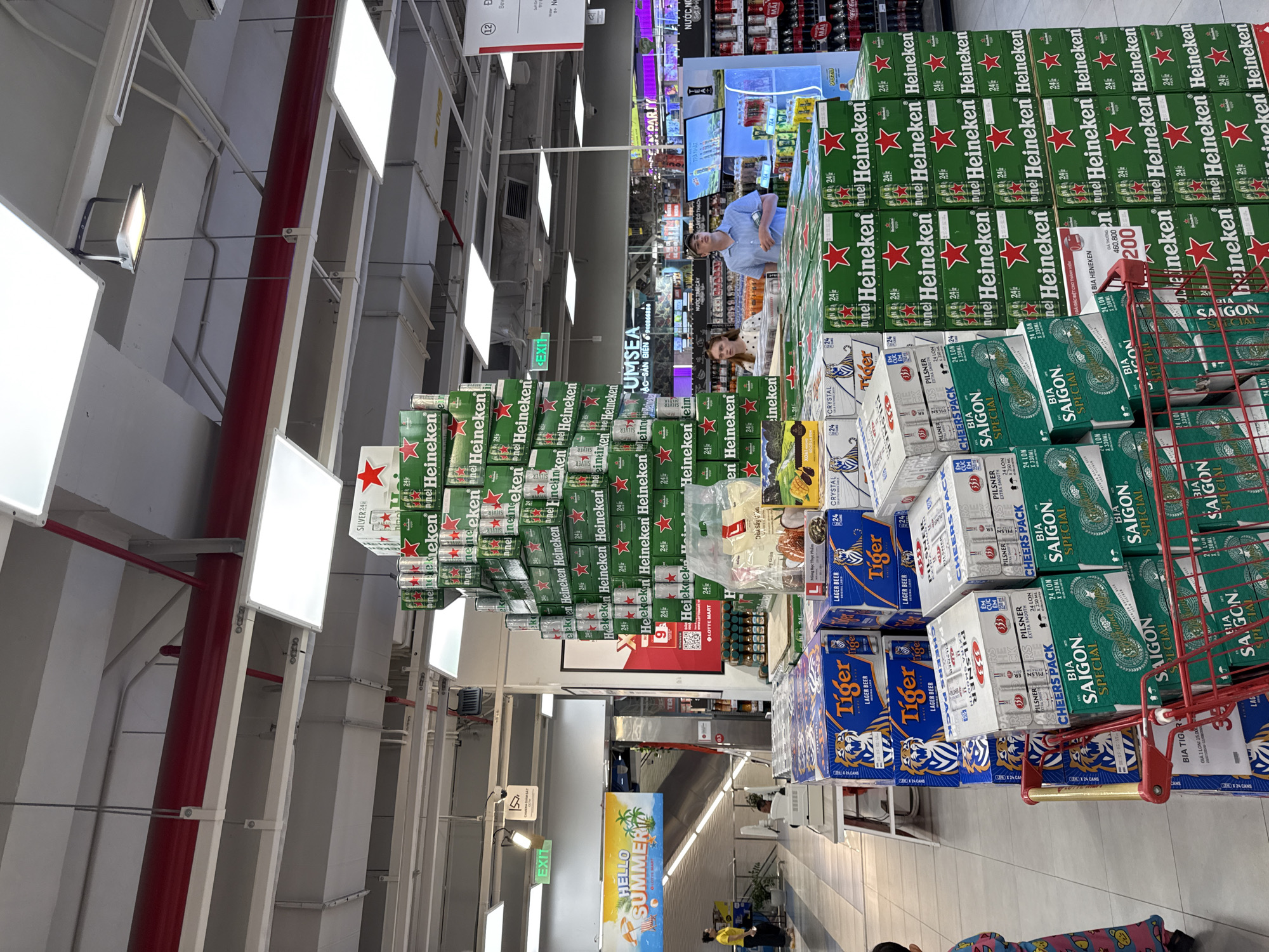 The image depicts the beverage aisle of a large supermarket in Nha Trang, Vietnam, as indicated by the "LOTTE MART" signage. Dominating the scene is a tall, pyramid-like display of green Heineken beer cases, stacked several layers high. To the left, there are stacks of "Tiger" beer boxes and "Saigon Special" beer cases. In the foreground, a red shopping cart is partially visible, filled with more beverage boxes. Two young adults, a male and a female, are standing in the middle ground, looking towards the display. In the background, there are shelves stocked with various drinks, overhead fluorescent lighting, and other store fixtures. A digital screen displaying advertisements is also visible on the right. The overall environment suggests a typical retail setting within a commercial establishment.