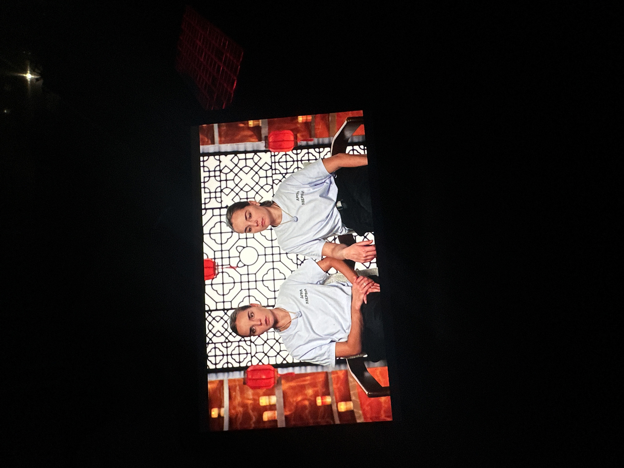 The image shows two young women, likely in their early twenties, seated in chairs on a dimly lit stage. The backdrop features a stylized, geometric pattern with red lanterns hanging from it, suggesting an East Asian theme. Both women are wearing light blue t-shirts with what appears to be text in Cyrillic script printed on the left breast, and black pants. They are both looking forward, with the woman on the left appearing to be speaking. The overall setting is likely a television studio or performance venue. There is no information in the image to suggest the location is Novoaltaysk, Russia.