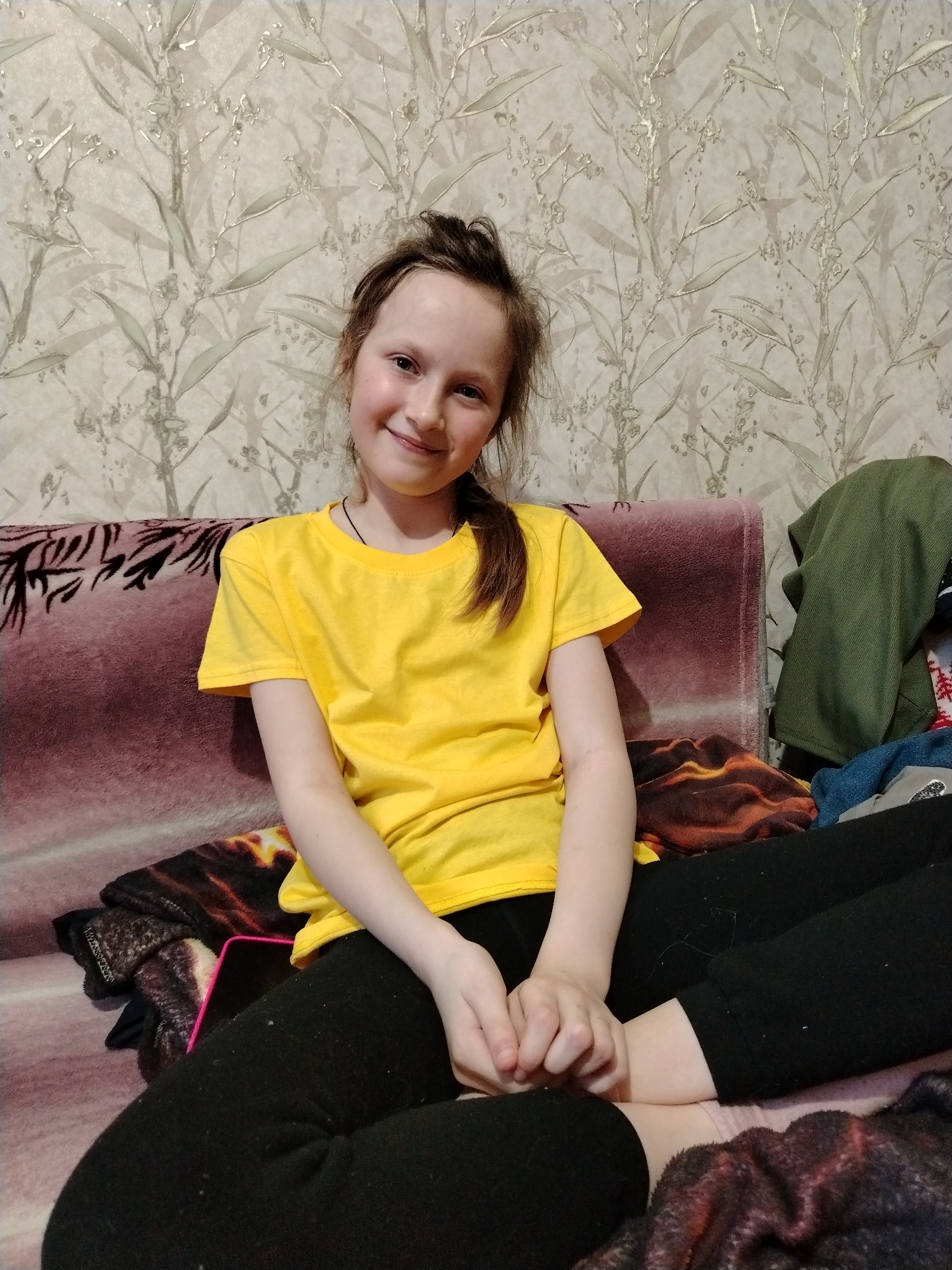 In an indoor setting in Nizhnyaya Gar', Russia, a young female is seated on a sofa, facing the viewer with a smile. She has a light complexion and brown hair tied into a ponytail. She is wearing a plain yellow short-sleeved t-shirt and black pants, with a thin black necklace around her neck. Her hands are clasped in her lap. The sofa features a faded reddish-purple upholstery, and a dark, multi-toned (brown, orange, black) patterned blanket is draped over her lower body and on the sofa beside her. To the subject's right, a folded olive-green garment and other clothing items are present on the sofa. A black smartphone with a pink border is partially visible beneath the patterned blanket near her left thigh. The wall behind her is covered with light-toned wallpaper depicting a repeating pattern of light green and silver-hued botanical elements.