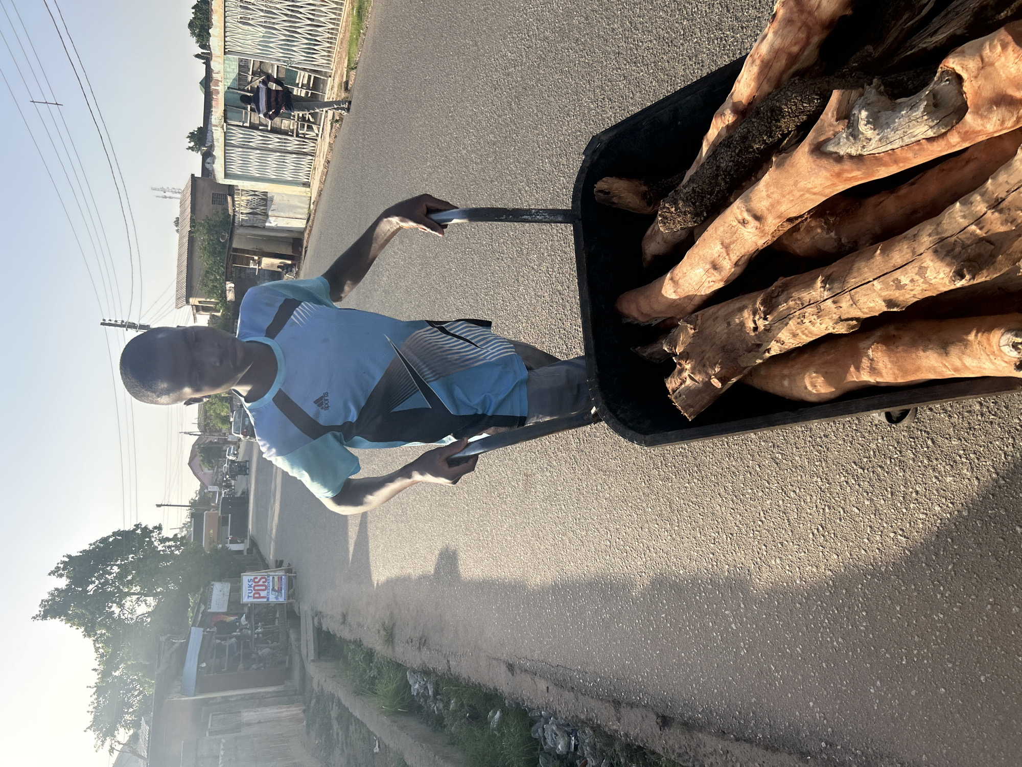 A man pulls a wheelbarrow filled with logs on a paved street in Jos, Nigeria. He is wearing a blue shirt and black shorts. The wheelbarrow is black and full of light brown logs. There are buildings and a fence visible in the background, along with other people. The photo appears to be taken during daylight hours with bright sunlight.