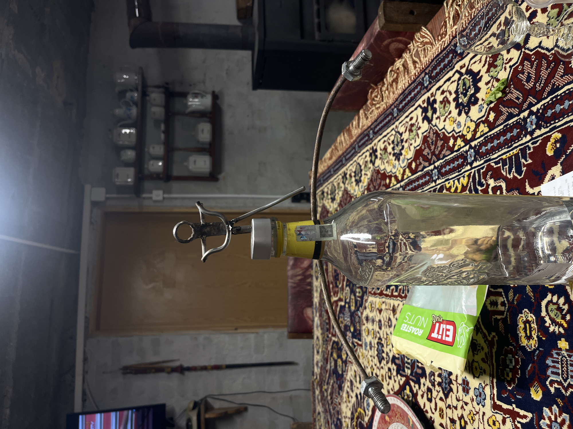 The image shows a cluttered indoor scene. In the foreground, a glass bottle filled with a clear liquid is held in place by a metal contraption. A bag of "ELIT ROASTED NUTS" rests on the bottle. The scene is set atop a patterned rug or tapestry.
In the background, a wall features shelves holding various white objects, and there's a wood-paneled door or wall section. A metal pipe extends from above. Below the door, there is a television and a long, thin decorative item that could be a sword or similar object.
The location, Lozarevo, Bulgaria, cannot be verified from the image.
