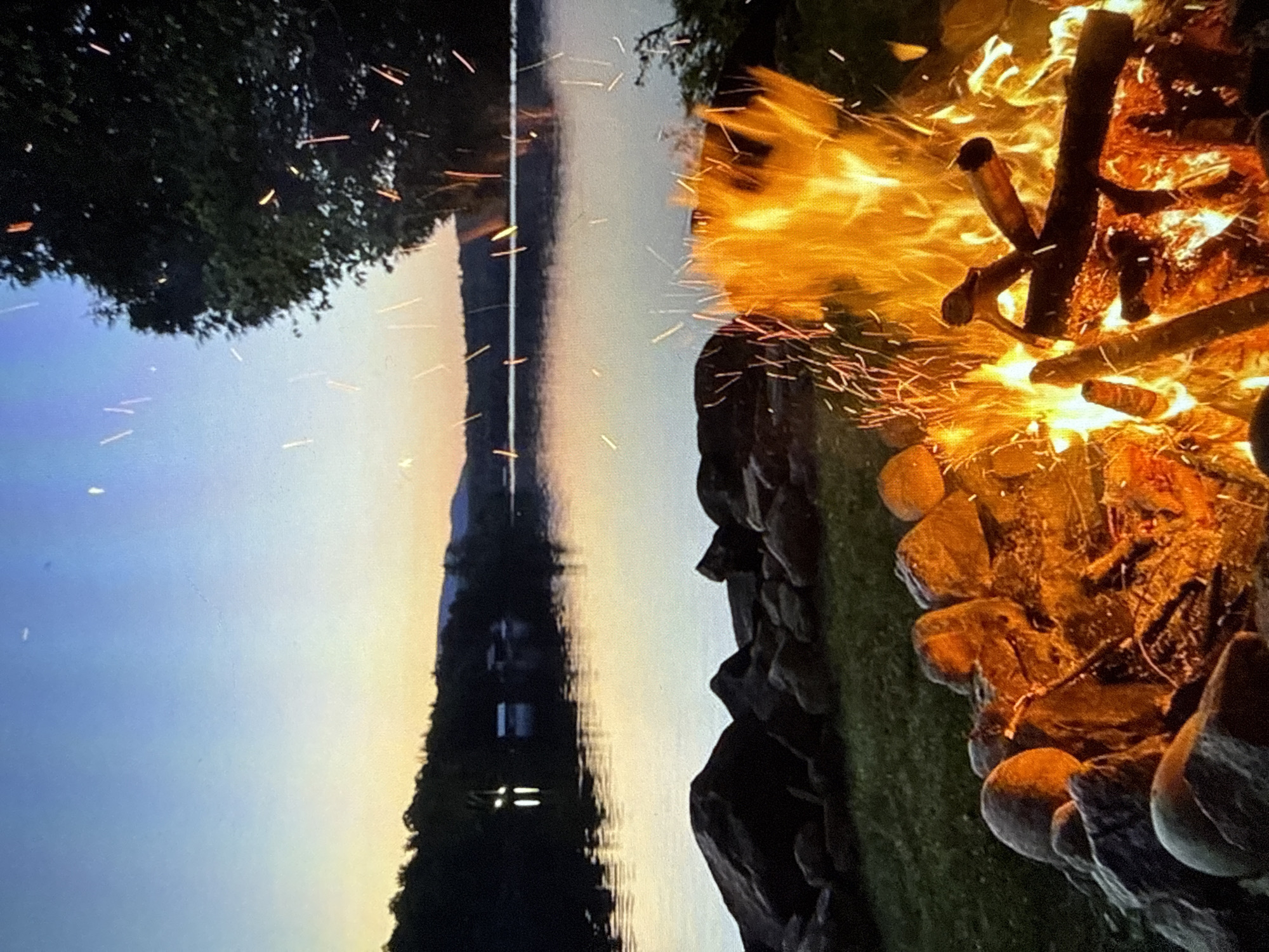 The image depicts an outdoor scene at dusk with a campfire burning in the foreground. The campfire is surrounded by stones and logs, with bright orange and yellow flames and sparks rising into the air. Behind the campfire, a body of water, likely a lake or a large river, reflects the soft colors of the sunset. A silhouetted treeline borders the water, and in the distance, faint mountain shapes can be seen against the twilight sky. The sky transitions from a pale blue at the top to a warm orange and yellow hue near the horizon. The overall atmosphere is serene and peaceful, suggesting a natural setting rather than an urban environment. There are no discernible elements that place this scene specifically within the city of Varna, Bulgaria.