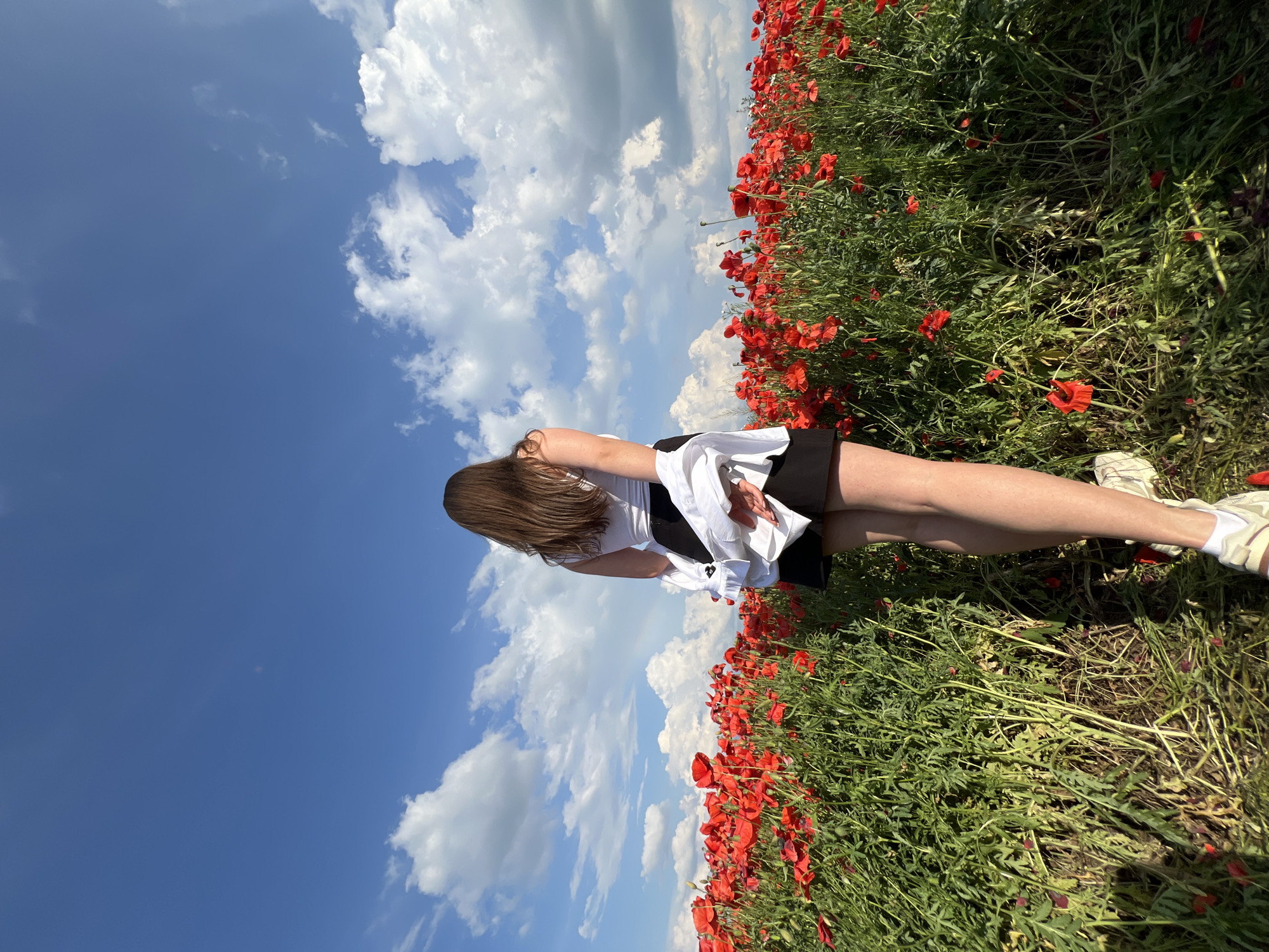 A young woman stands in a field of red poppies, facing away from the camera. She has long, dark brown hair and is wearing a white tank top, a black skirt, and a white shirt draped over her shoulders. The field of poppies extends into the distance, under a bright blue sky dotted with fluffy white clouds. The scene appears to be a natural landscape, likely a meadow or countryside, and does not contain any elements that would suggest a specific location within the city of Ternopil, Ukraine, or any urban setting.