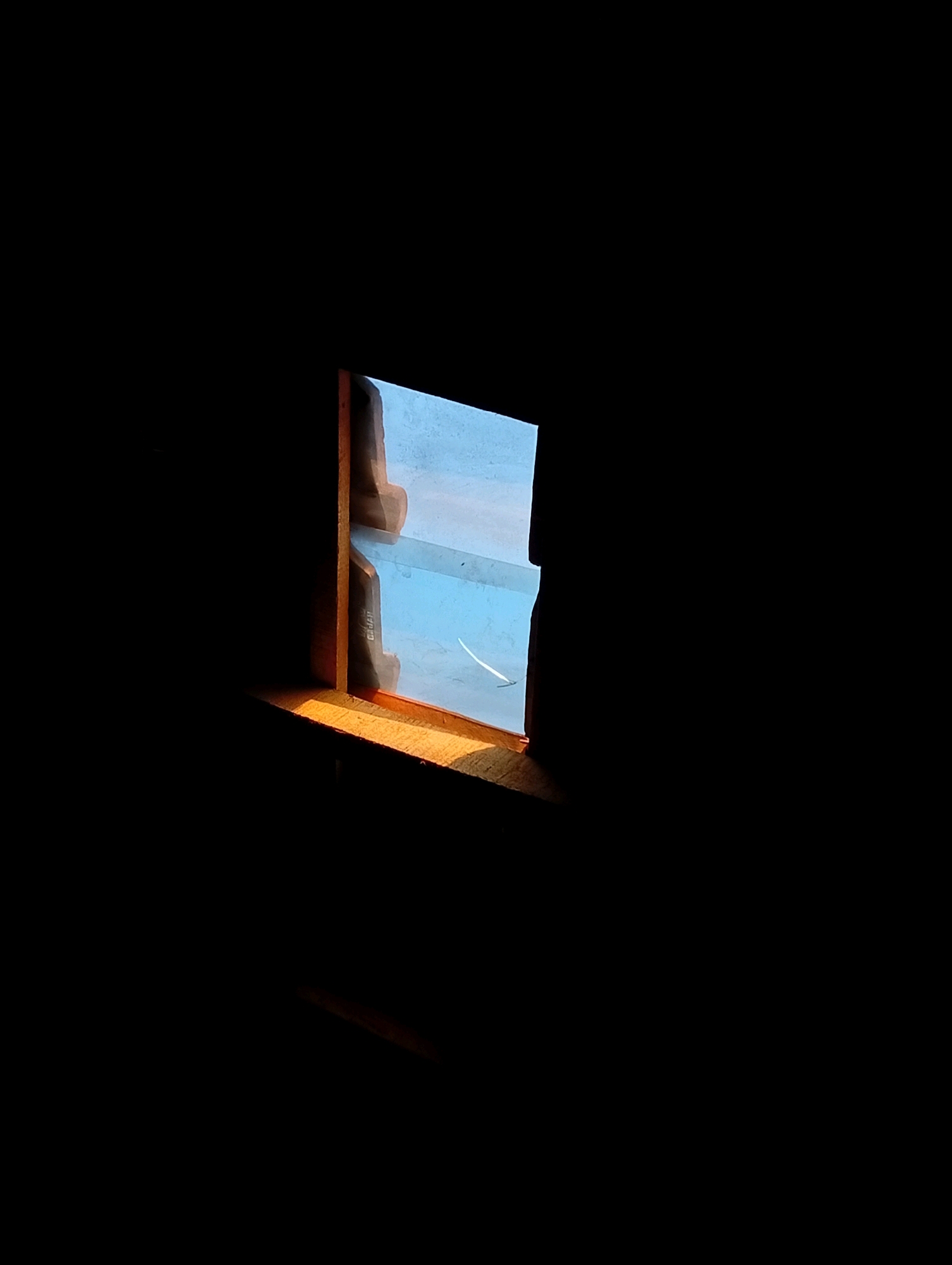 The image shows a small, partially open window, with wooden frames, set against a very dark, almost black background. Sunlight streams through the window, illuminating the wooden sill with a warm, orange glow. The glass panes of the window are a pale blue, reflecting the light and creating a bright, airy contrast to the surrounding darkness. Through the window, a blurred, abstract view of the outside is visible, appearing as soft shapes and colors. The overall impression is one of quiet solitude, with the light acting as a focal point within the deep shadow. There is no information about the city of Kebandingan, Indonesia in this image.