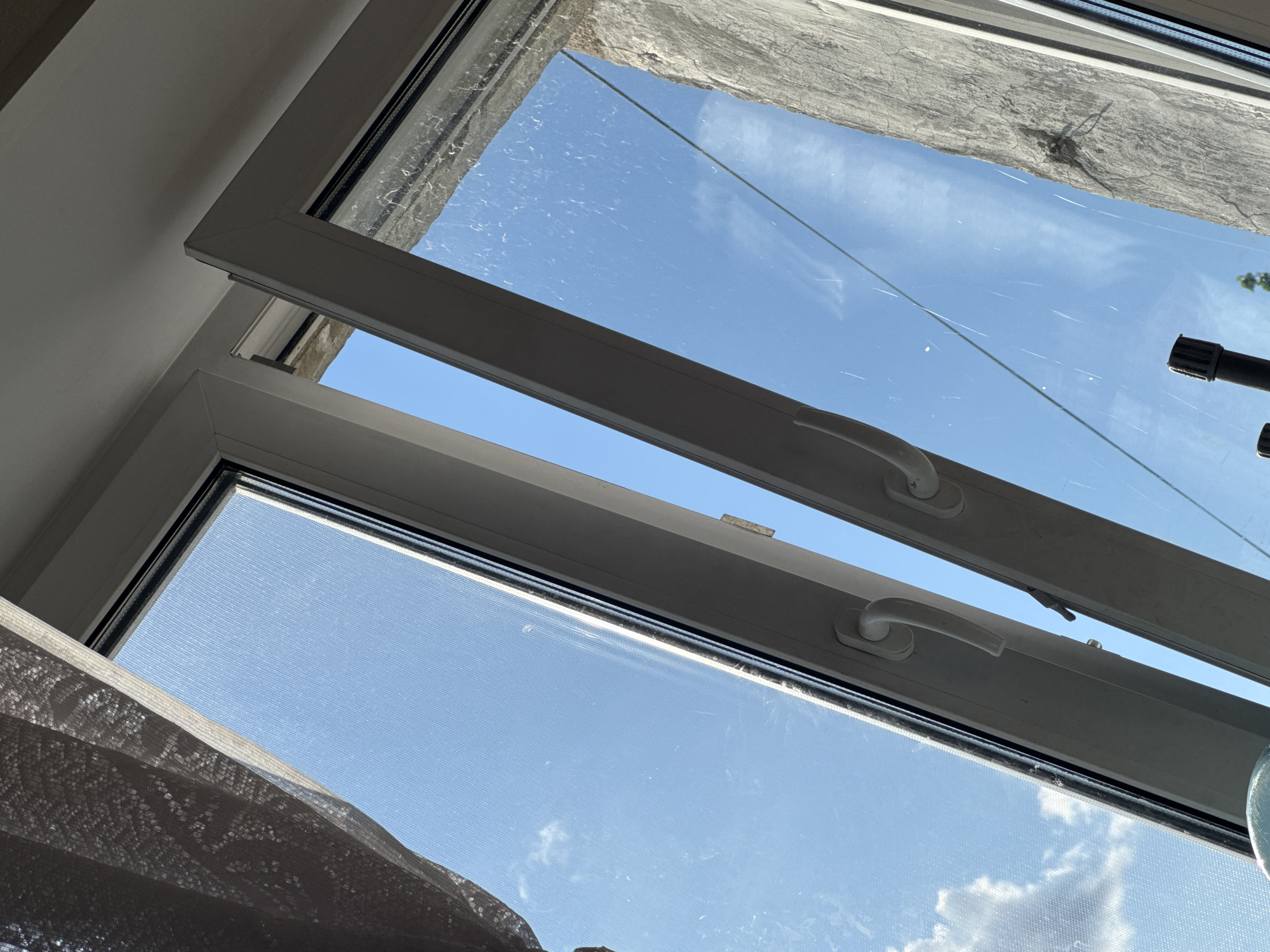 The image shows a close-up view of an open window, looking upwards towards a bright blue sky with scattered white clouds. The window frame is white PVC. On the left side of the frame, a textured light-colored curtain is partially visible. The window pane on the left appears to have a fine mesh screen. The window on the right is open, revealing a rough, textured concrete wall outside. There are two white window handles visible, one on each section of the window. A black object, possibly a flashlight or part of a tool, is visible in the lower right portion of the frame. The scene appears to be outdoors, likely on a sunny day. There is no identifiable information in the image to indicate the specific location within the city of Chelyabinsk, Russia.