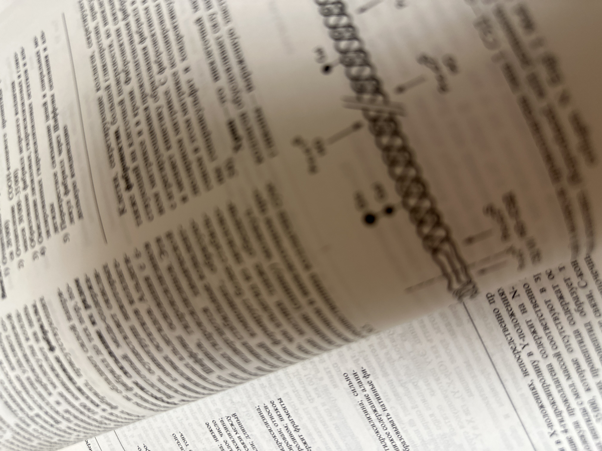 The image shows a close-up of an open book with Russian text. The book appears to be a scientific or medical textbook, with dense paragraphs and some smaller text entries. A diagram is partially visible on the right side of the image, depicting a molecular structure with labels. The lighting suggests the photograph was taken indoors, possibly in a library or study. There are no people visible in the image. The focus is on the text and diagram, with some areas of the page being blurred due to the camera's depth of field. The book is lying flat on a surface, and the angle of the shot is from above and to the side.