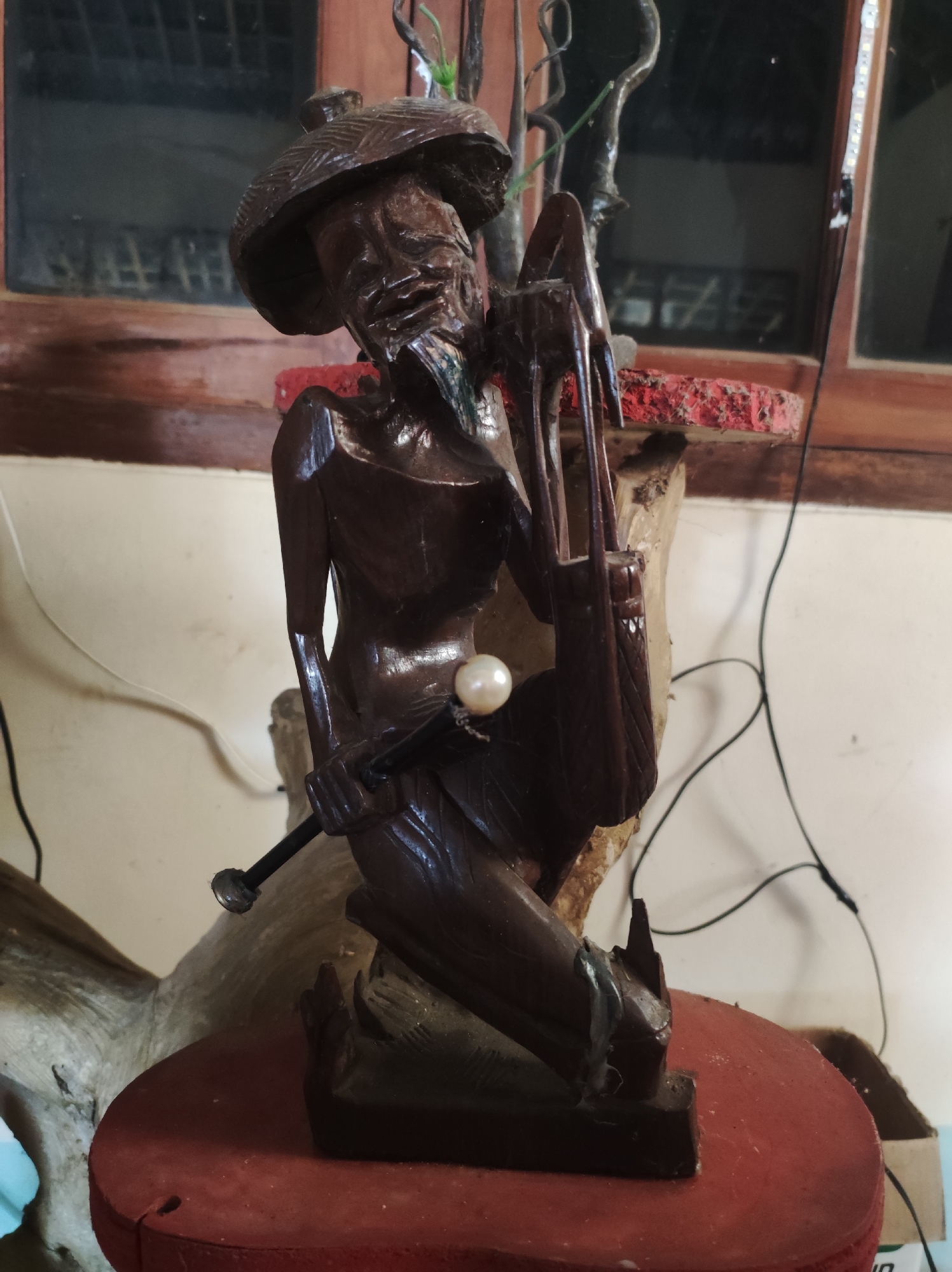 A dark brown wooden sculpture of an elderly man sits on a red circular base. The man is wearing a conical hat and a garment that drapes over his body. He is holding a staff topped with a pearl in his right hand. His left hand rests on a bag-like object. The sculpture appears to be placed indoors, with a wooden window frame and what looks like a red painted wooden platform in the background. Electrical wires are visible in the scene. The overall impression is that of an indoor decorative piece. The location is specified as Mojopuro, Indonesia.