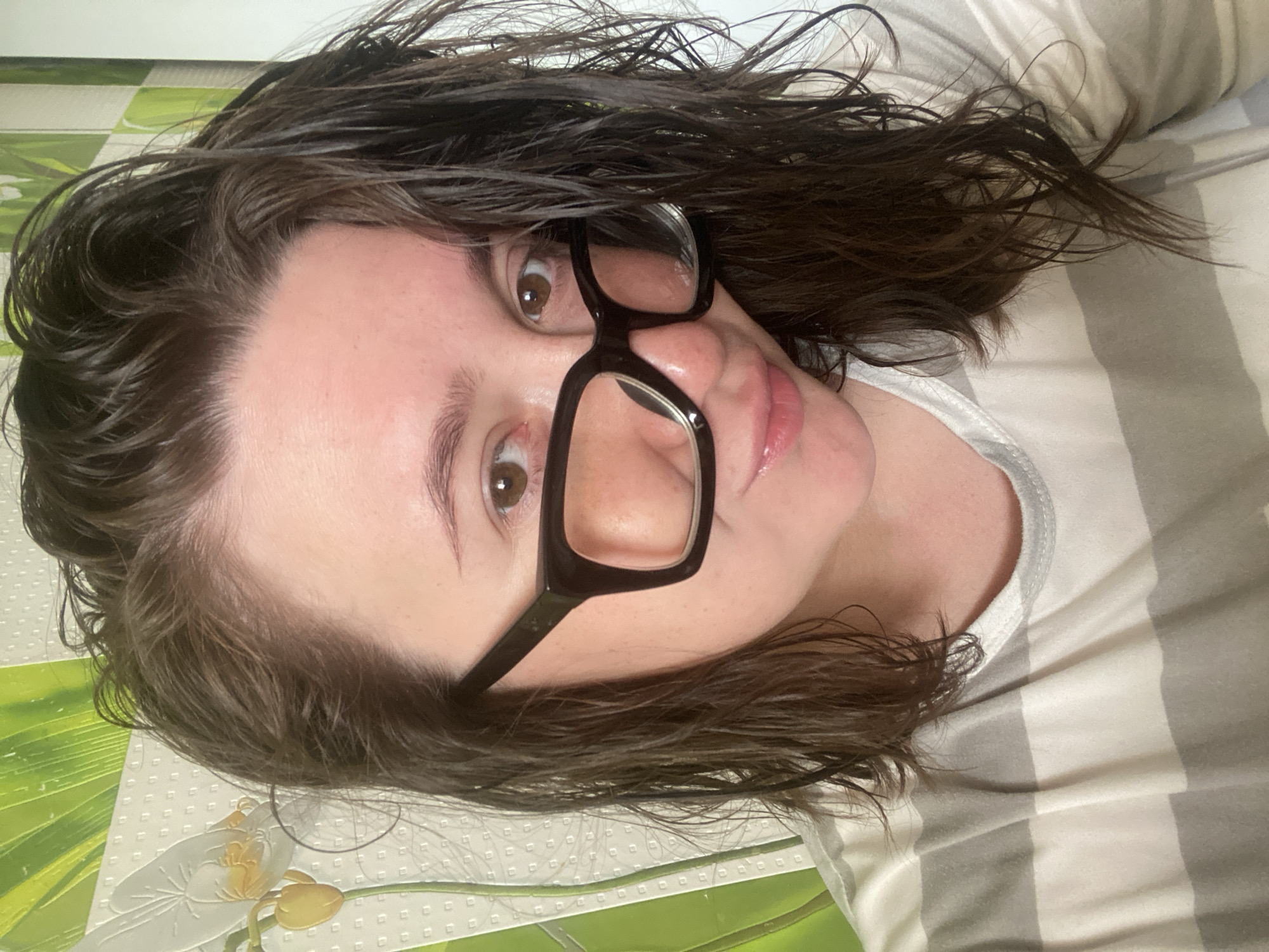 The image depicts a medium close-up portrait of an adult female, reportedly located in the city of Chaikovsky, Russia, though no specific geographical markers within the photograph confirm this location. The subject has wet, dark brown hair, extending to her shoulders, which appears somewhat disheveled. She possesses fair skin, brown eyes, and full lips with a subtle sheen. She is wearing black, thick-rimmed, rectangular glasses and looks directly into the camera with a slight, gentle smile. Her attire consists of a crew-neck t-shirt patterned with horizontal stripes in alternating light gray and off-white. The background features a light-colored wall covering with a repeating grid pattern, overlaid with stylized green foliage and a prominent white flower, possibly an orchid, detailed with yellow accents. The lighting is bright and appears even.