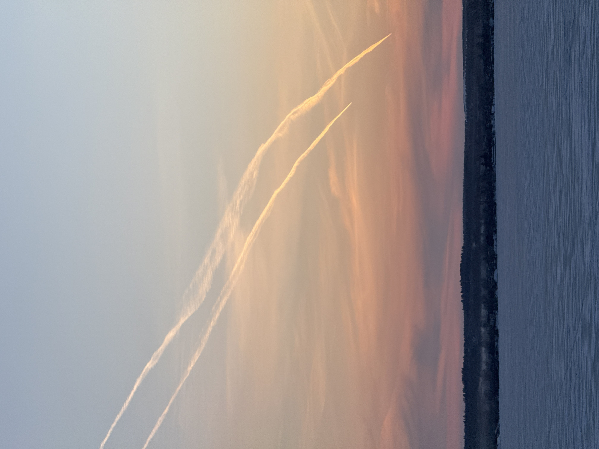 The image depicts a sunset over a body of water, presumably a lake or river, with a shoreline visible in the distance. Two contrails are visible in the sky. The sky is a gradient of colors, ranging from blue to orange to pink. The water is a dark blue. The shoreline is marked by a line of trees. Given the context, this may be Votkinsk, Russia, and the water body could be the Votkinsk Reservoir.