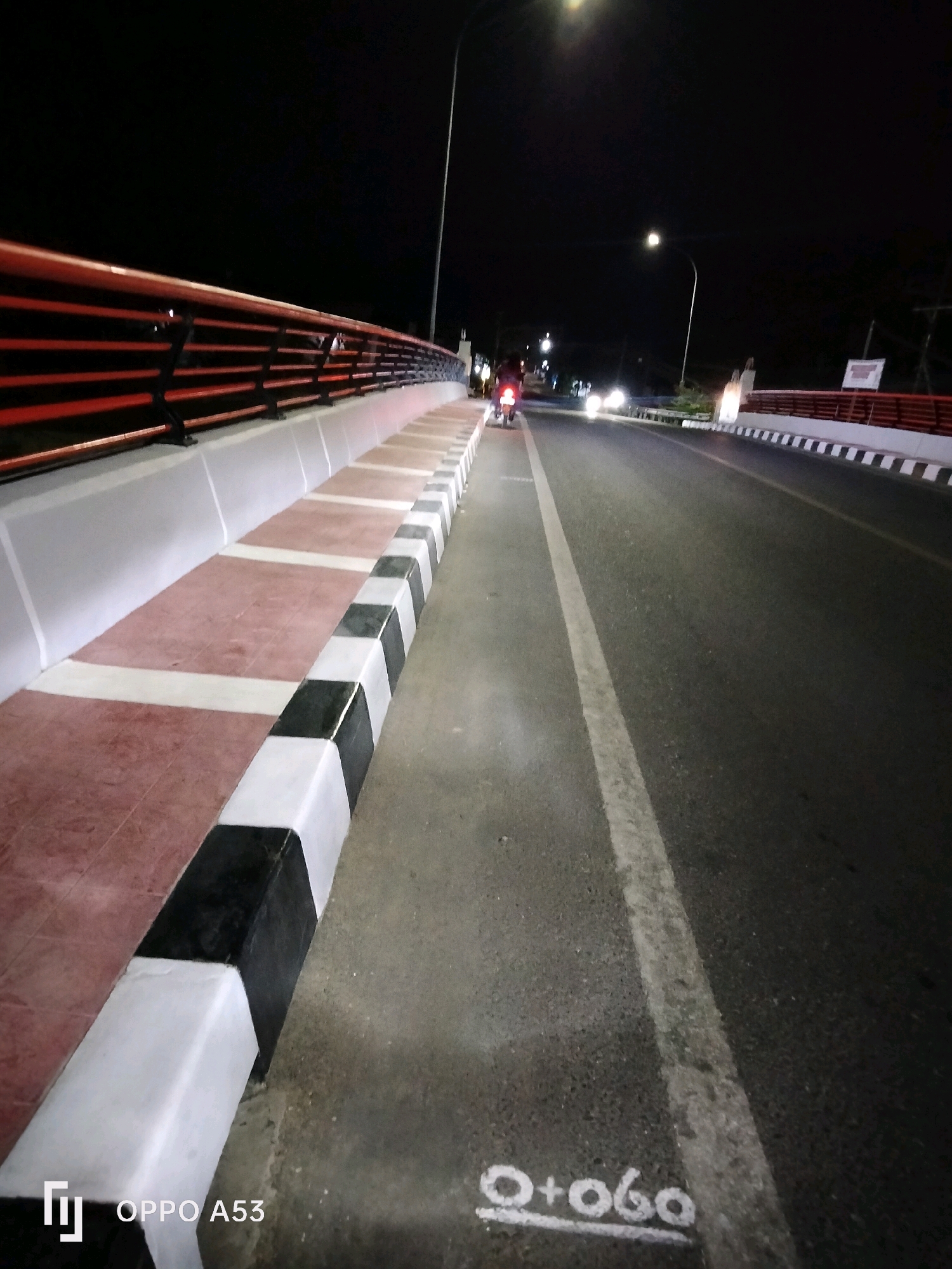 A night view of a bridge in Bendo, Indonesia. The bridge has a red railing and white concrete barriers with black and white stripes. There is a sidewalk on the left side of the road with reddish tiles and white and black checkered barriers. The road is dark asphalt with a white line marking the lane. In the distance, there are streetlights and car headlights. One motorcycle with a rider is visible on the road. There are some markings on the road that say "O+060" with a line underneath. The OPPO A53 logo is visible in the bottom left corner.