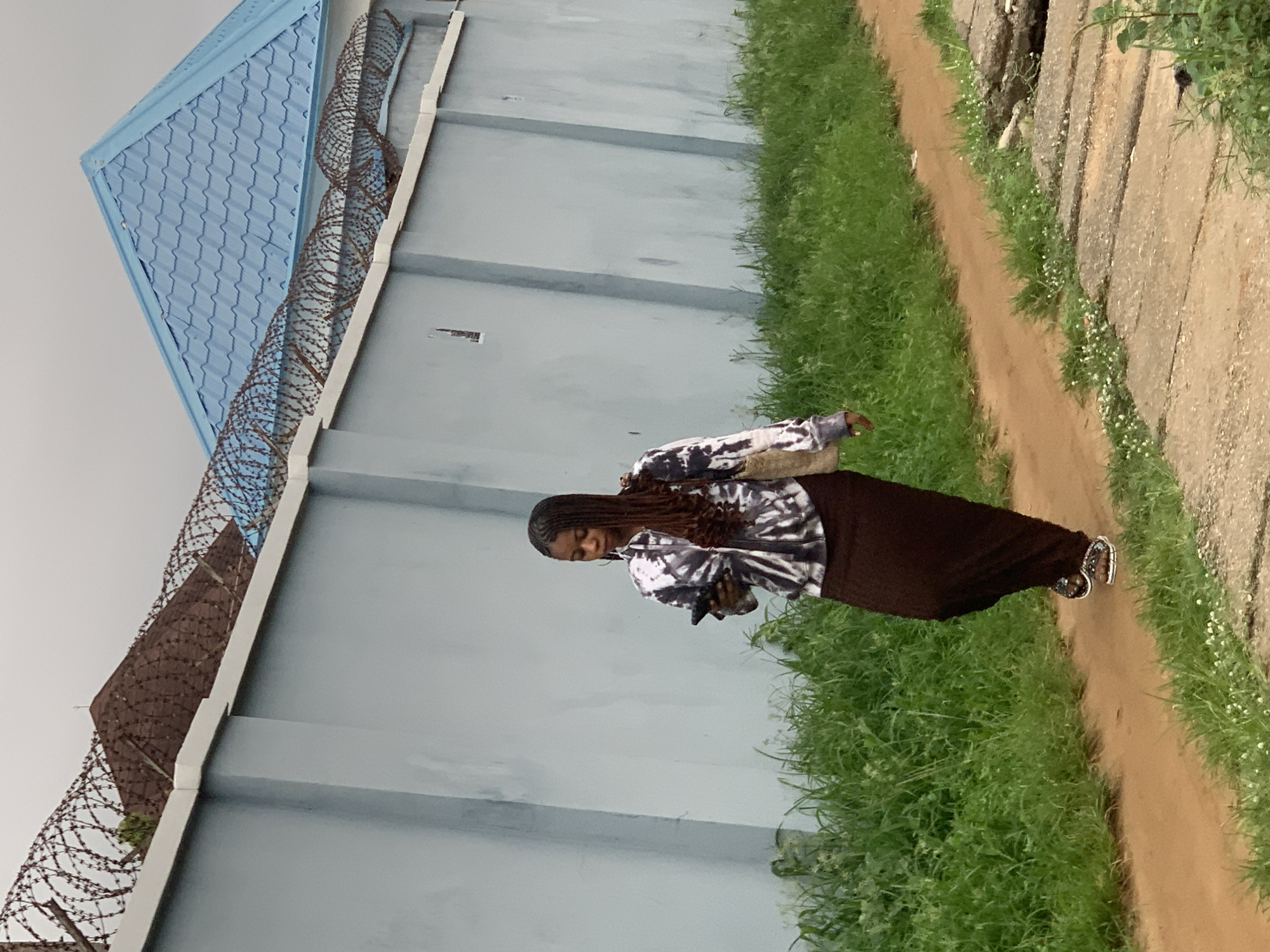 A young woman with long, braided hair is walking on a dirt path beside a light blue concrete wall. She is wearing a tie-dye jacket and a dark brown, floor-length skirt. She is holding a black mobile phone in her right hand and has a light-colored woven bag slung over her left shoulder. The path is bordered by lush green grass on both sides. Behind the wall, a portion of a house with a brown, tiled roof and a section of a light blue, corrugated metal roof are visible, topped with barbed wire. The sky is overcast. The scene is located in Abuja, Nigeria.