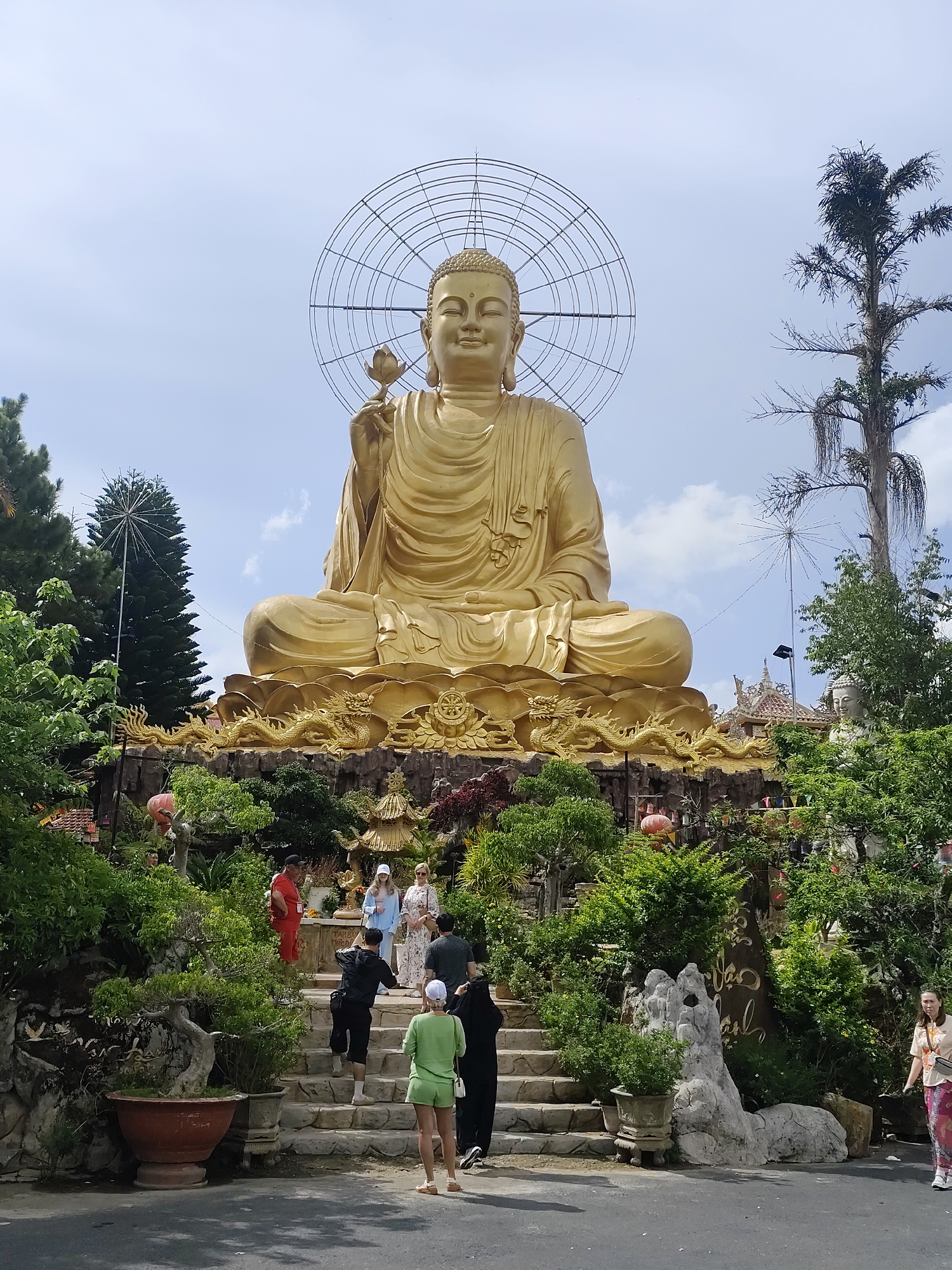 This photograph captures a large golden Buddha statue seated atop an elevated platform surrounded by lush greenery in Lâm Viên - Đà Lạt, Vietnam. The Buddha is in a meditative pose, holding a lotus flower, with a circular halo-like structure behind its head. Several people are walking up steps towards the statue, including a person in a green outfit. The scene is set against a partly cloudy sky. The surrounding garden area features a variety of plants, decorative stone features, and traditional Vietnamese architectural elements.