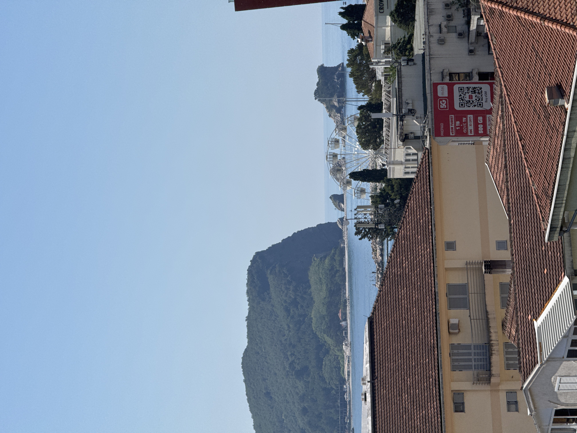 The image captures a coastal city scene, likely Budva, Montenegro, on a clear, sunny day. In the foreground, terracotta tiled roofs of buildings are prominent, with one building featuring a pale yellow facade, a balcony with a metal railing, and windows. Further back, more buildings with white and beige exteriors are visible, some equipped with air conditioning units and satellite dishes.
The middle ground reveals a Ferris wheel situated near the water, with several rocky islets scattered in the blue sea. Beyond the islets, a large, tree-covered hill or mountain dominates the background, meeting the horizon with the clear sky. In the distance, a sailboat can be seen on the water. A red advertisement sign for "5G m:tel" is prominently displayed on one of the buildings, offering various data packages and the option to "Buy eSIM." The scene appears to be a typical urban and tourist landscape, blending residential and commercial structures with natural coastal elements. There are no people or animals clearly visible in the frame.