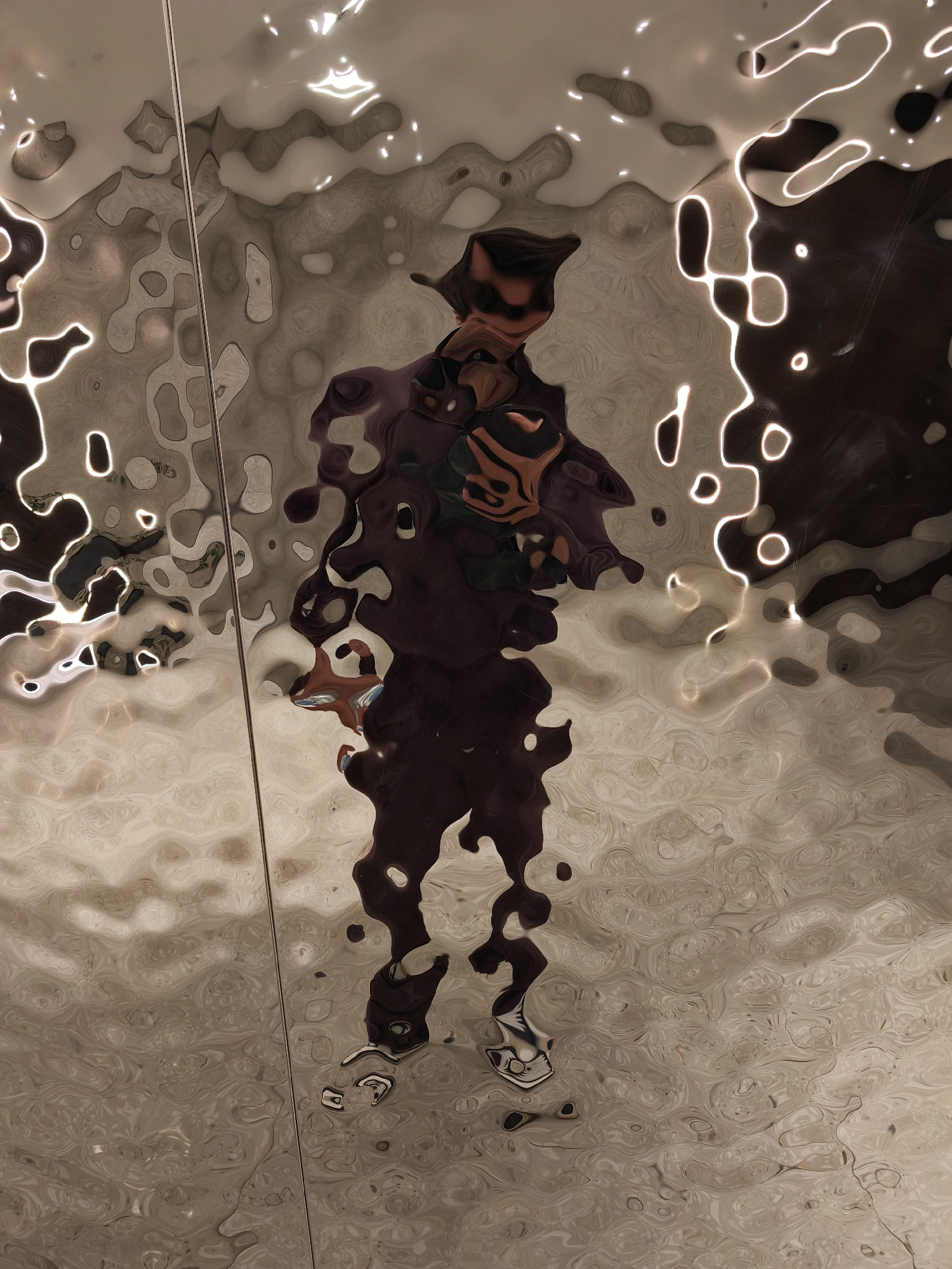 The image shows the distorted reflection of a person in a textured, reflective surface. The person is standing and appears to be wearing a hat or head covering. The reflection is stretched and warped due to the uneven surface. The background is a mix of light and dark patterns. The location appears to be Moscow, Russia, based on the appearance of the reflective surface, which seems to be part of a public art installation or architectural feature.