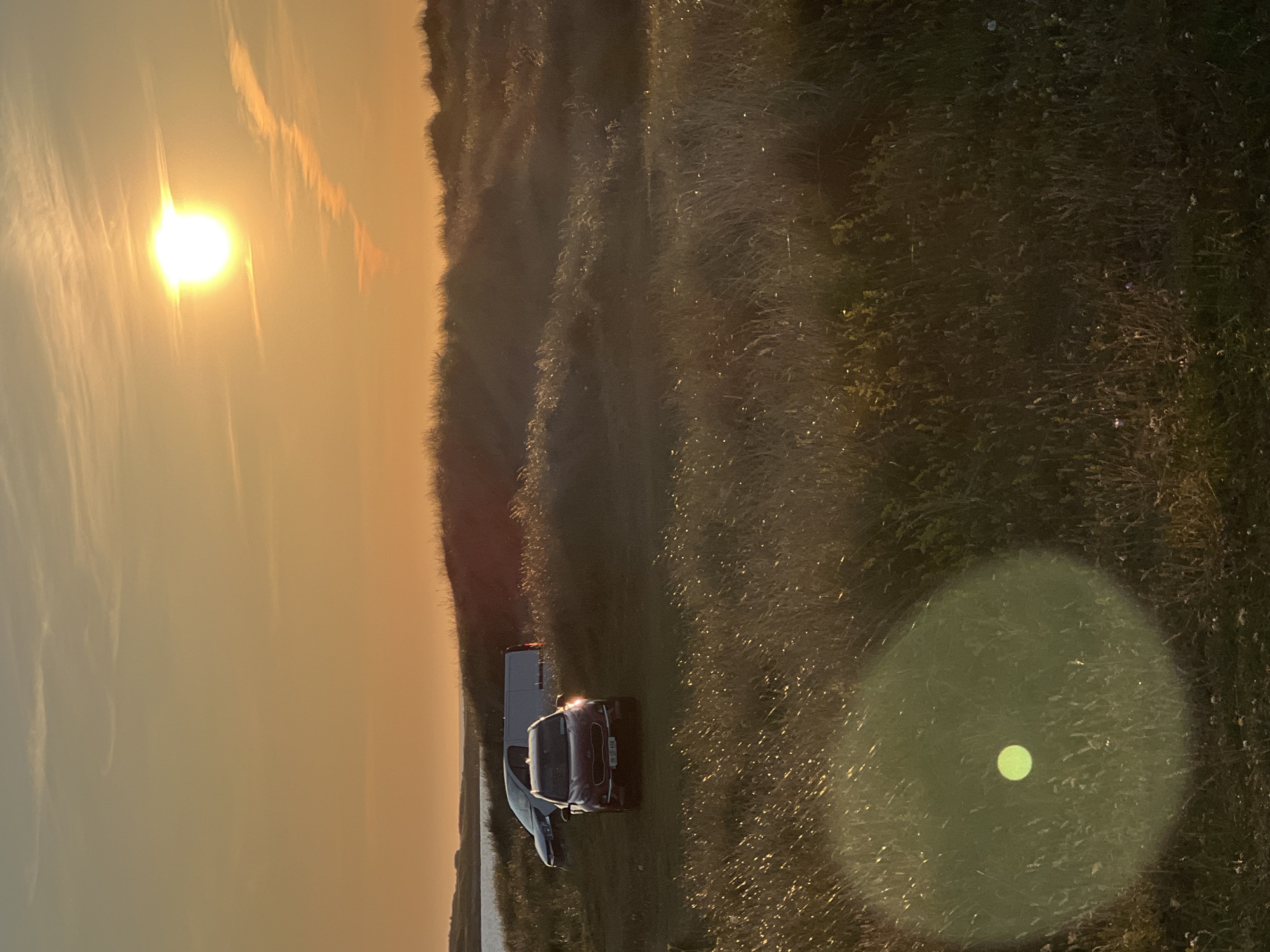 The image depicts a coastal landscape at sunset in Stranacorkra, Ireland. The sky is a warm orange and yellow gradient, with a bright sun positioned in the upper right quadrant. Wispy clouds are visible, streaking across the sky. The foreground is dominated by tall, dry grass, which is illuminated by the setting sun, creating a shimmering effect. A large, circular lens flare is present in the lower left portion of the image.
In the middle ground, there are several vehicles parked on a grassy area. A dark red SUV is prominently featured in the center, with its headlights on. Behind it, a white van and a silver car are partially visible. The vehicles are situated amongst rolling dunes covered in vegetation. In the far distance, a body of water is visible, likely the Atlantic Ocean, with a low-lying coastline on the horizon. The overall scene suggests a serene, natural environment bathed in the warm light of sunset.