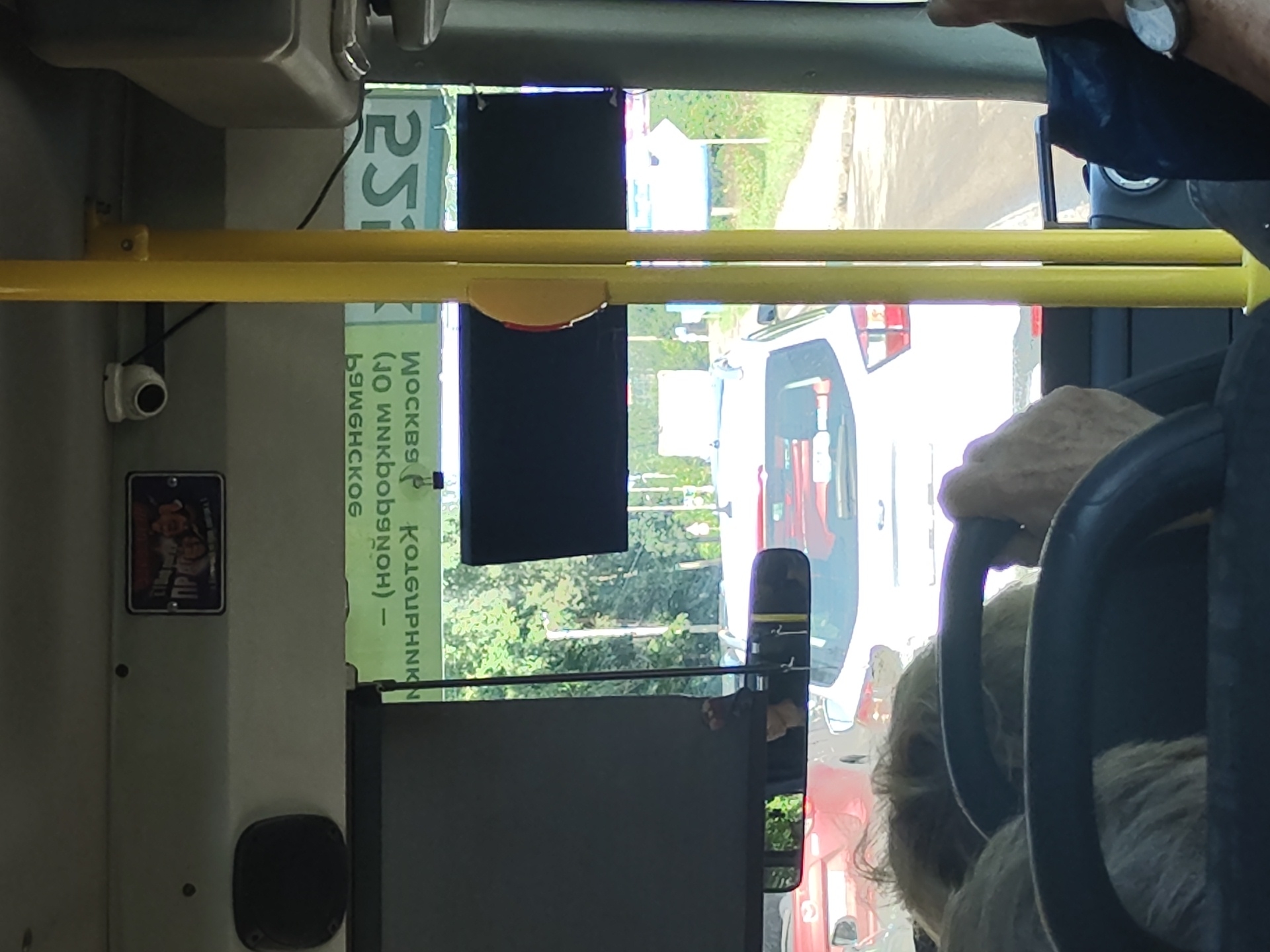 The image shows the interior of a bus with a view out the front windshield. A yellow pole runs vertically through the center of the frame. Through the windshield, traffic is visible, including a white car and red cars. In the distance, trees and a sidewalk can be seen. On the dashboard, there is a small sticker with an image and Cyrillic text. Above the windshield, a security camera is mounted. On the side of the bus, a sign with Cyrillic text is visible, which appears to be a route indicator. Based on the text on the sign which reads "Раменское – (ноябрь) 90. МОСКВА", the location is Ramenskoye, Russia, likely indicating a bus route to or from Moscow. The image is taken from the perspective of a passenger or the driver, as the steering wheel and part of the dashboard are visible in the foreground.