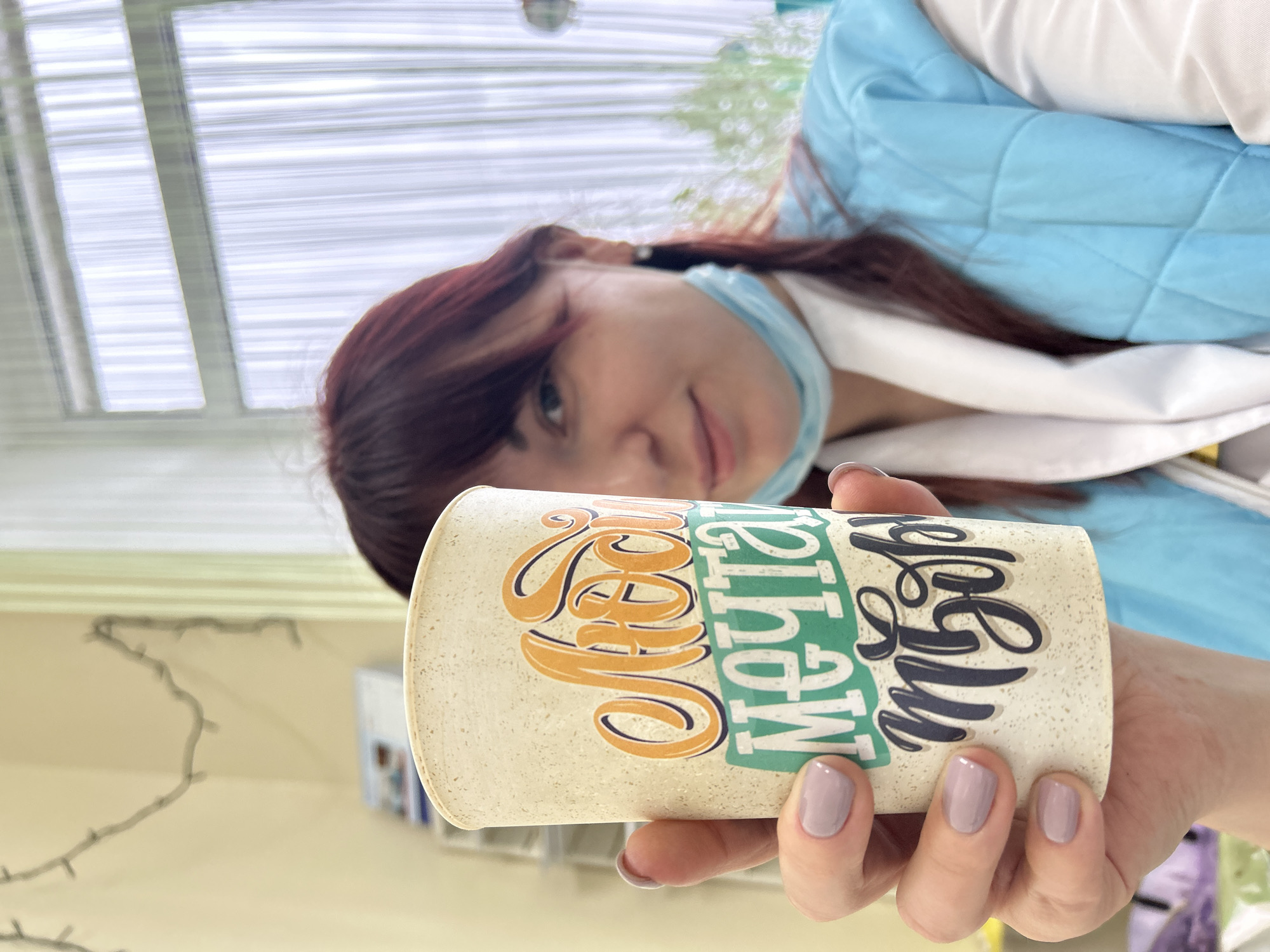 A woman with auburn hair, a blue surgical mask pulled down around her neck, and a light blue quilted jacket is holding a cylindrical cup with beige background, green letters that read "slow" and blue letters "life". Her fingernails are painted mauve. Behind her is a window with horizontal blinds, and string lights draped across a light yellow wall. Based on the location, this image was taken in Фоки, Russia.