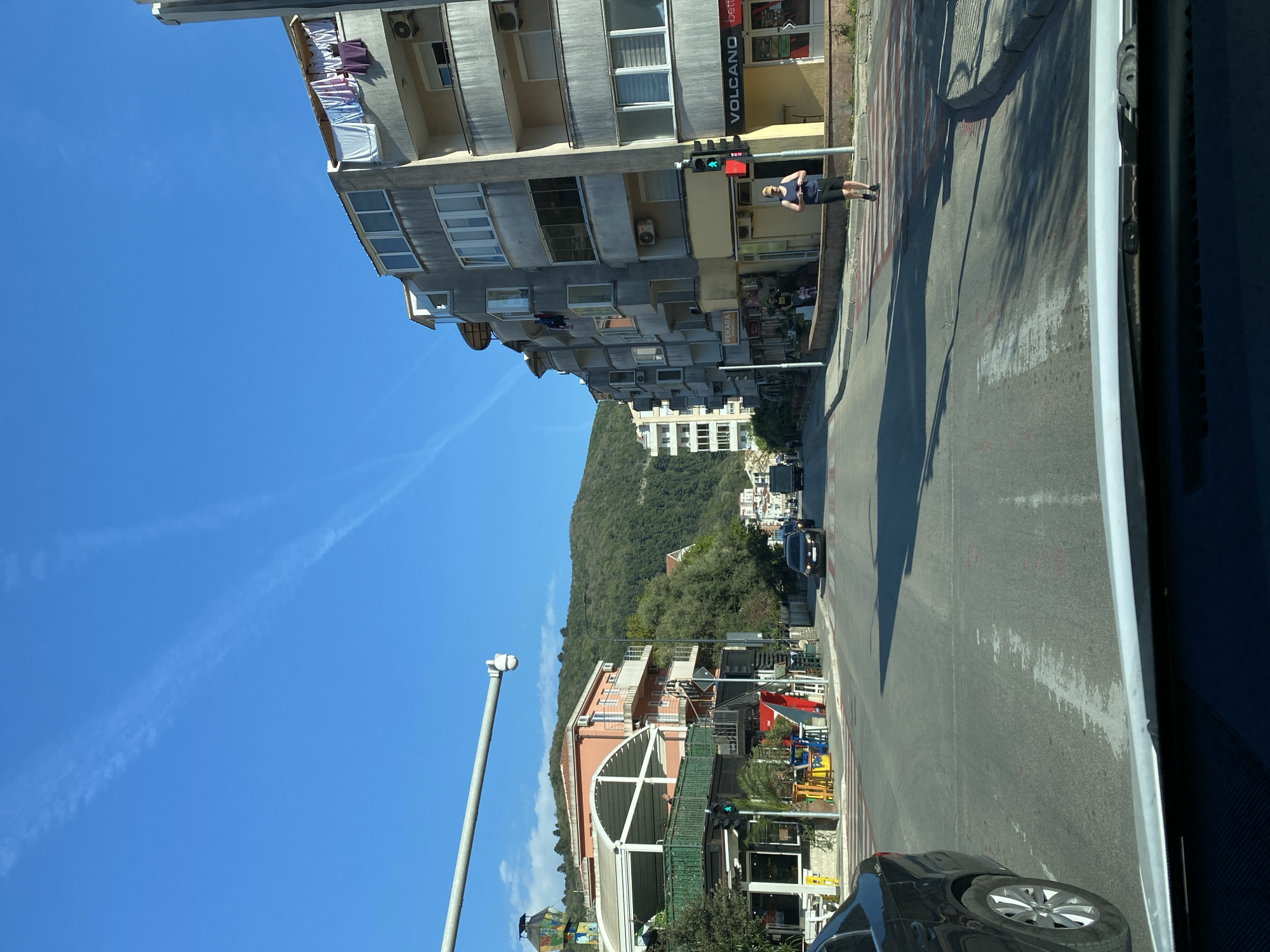 The image depicts a street scene in Budva, Montenegro, under a clear blue sky. The view is from inside a vehicle, looking across the road. A pedestrian stands at a crosswalk, waiting for the traffic signal to change. The signal is green for traffic moving along the road, and red for the crosswalk. The street is lined with buildings, including a multi-story apartment complex with balconies, a storefront labeled "VOLCANO" in large letters, and another business called "PEKARA" (Bakery). A car is partially visible in the foreground. Further up the street, there are more buildings, trees, and a hillside in the background. Contrails are visible in the clear blue sky.