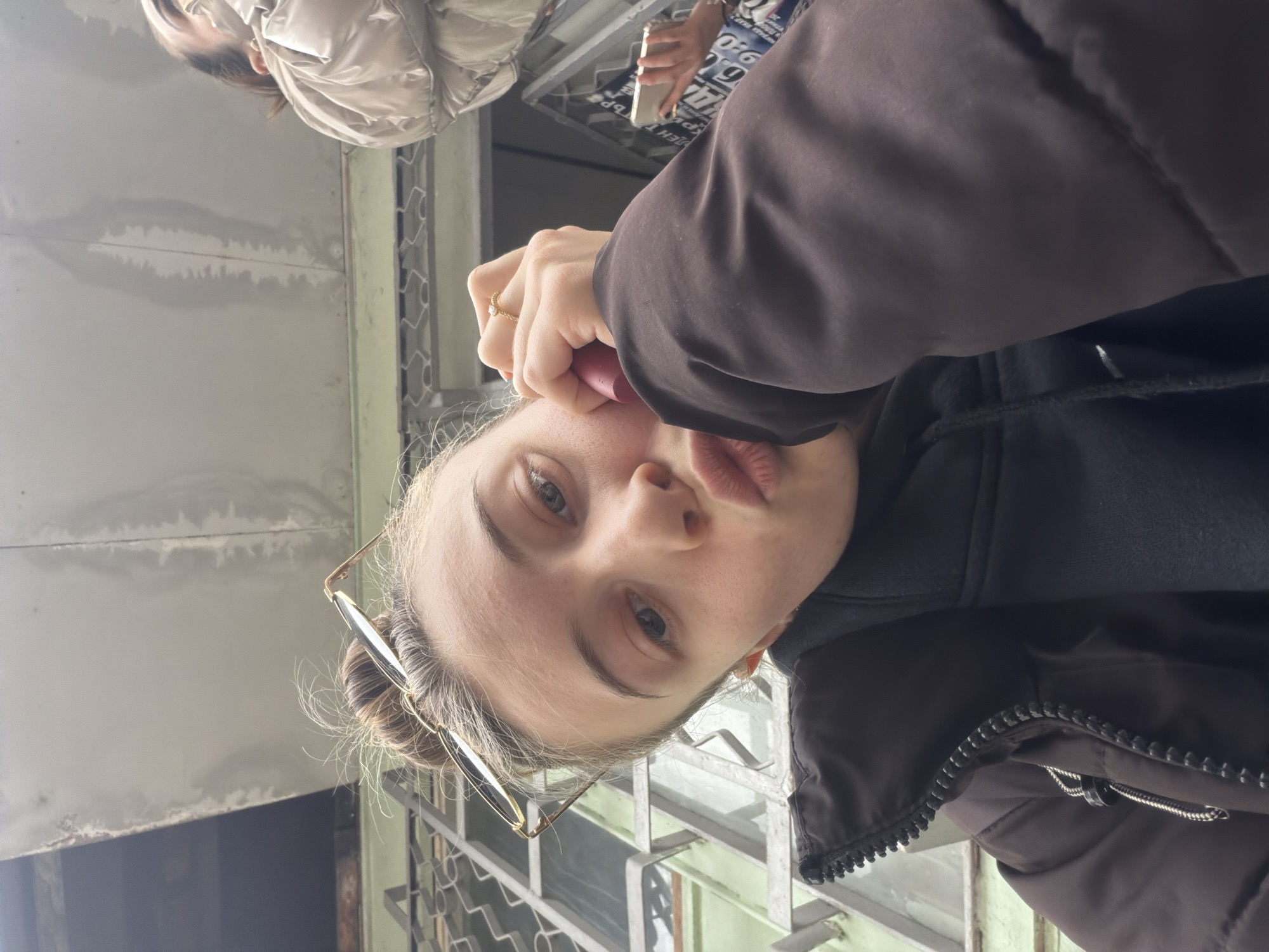 A woman with fair skin and light brown hair tied up and wearing glasses, a brown jacket over a black hoodie, is taking a selfie. She is holding a red lipstick tube in her right hand. Her expression involves pursed lips. Another person wearing a light-colored jacket is visible in the background, partially obscured. The selfie appears to be taken outdoors near a building with grey walls and window frames that suggest it might be a balcony or terrace. The building's architecture and color palette suggest a location within Plovdiv, Bulgaria. Another person is visible in the background, holding a cell phone.