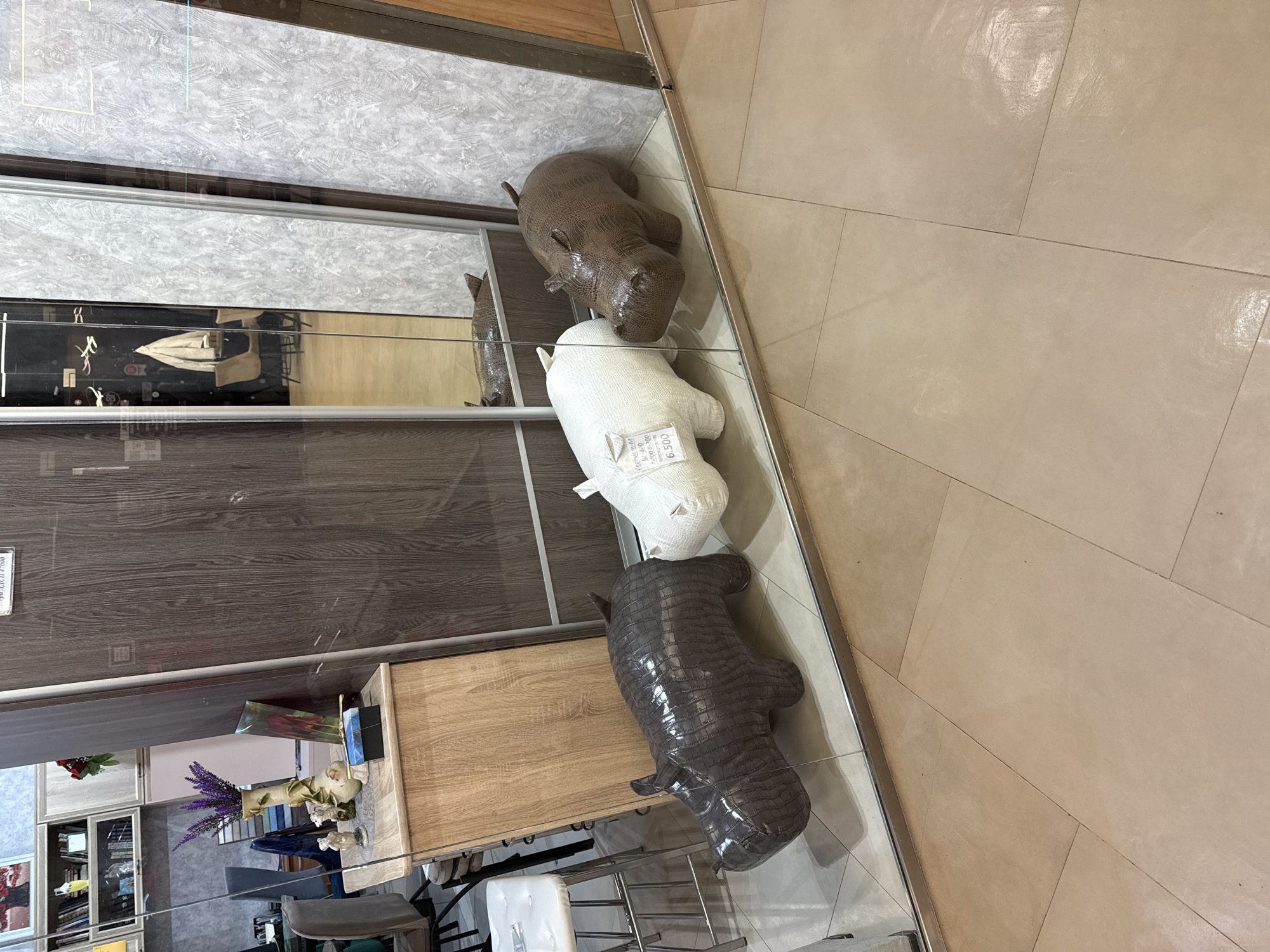 A ground-level, indoor shot depicts three hippo-shaped figurines of differing colors (brown, white, and gray-brown) arranged linearly along a metallic track, likely a part of a display unit or furniture set in a commercial space or home furnishing store. The figurines appear to be made of a fabric or soft material with a textured, possibly synthetic, skin. A tag is visible on the white hippo, implying a price.
The background showcases a reflection in the glass, revealing additional furniture pieces, decor items such as flowers, and a bookcase stocked with books and decorative items. The mirrored surface also shows reflections of the interior design elements.
The floor is covered with light tan-colored tiles, laid in a diagonal pattern. The angle of the shot and the tile pattern provides a sense of perspective, giving depth to the scene.
Given the details, it is not possible to verify the location of the image.