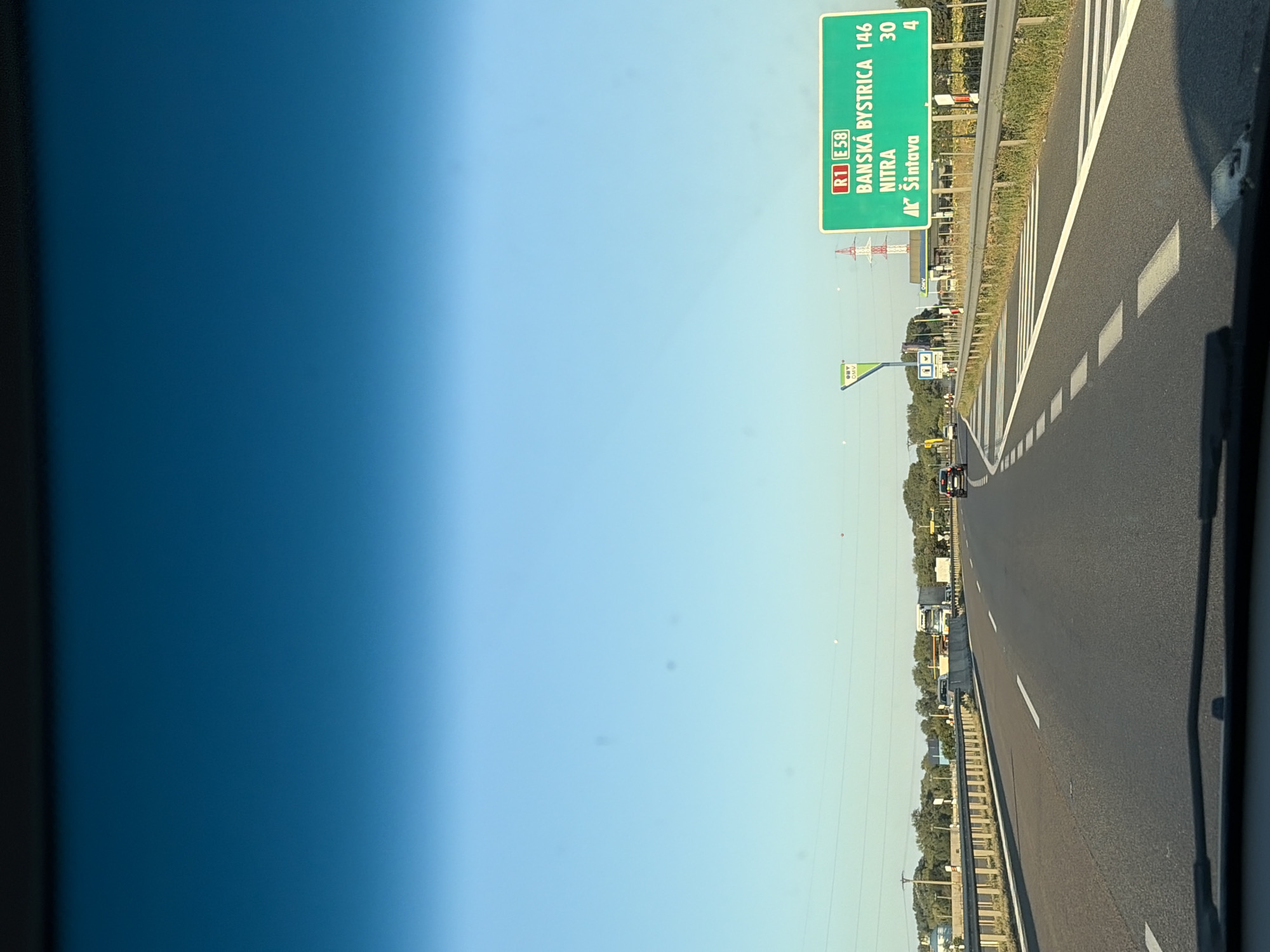 The image captures a view from the driver's perspective while on a highway, likely in or near Dolná Streda, Slovakia. The sky is a clear, bright blue, transitioning to a darker blue at the top of the frame, suggesting it might be early morning or late afternoon. A green highway sign is prominently visible on the right side of the road. This sign indicates directions to "BANSKÁ BYSTRICA" with a distance of 146, "NITRA" with a distance of 30, and "ŠINTAVA" with a distance of 4. Below the highway sign, a smaller green sign with the OMV logo is visible, indicating a gas station is nearby. The highway itself has multiple lanes and is bordered by a metal guardrail. Several vehicles, including cars and a truck, are present on the road. Trees and vegetation line the sides of the highway, and a power transmission tower can be seen in the distance. The road markings, including white dashed lines, are clearly visible.