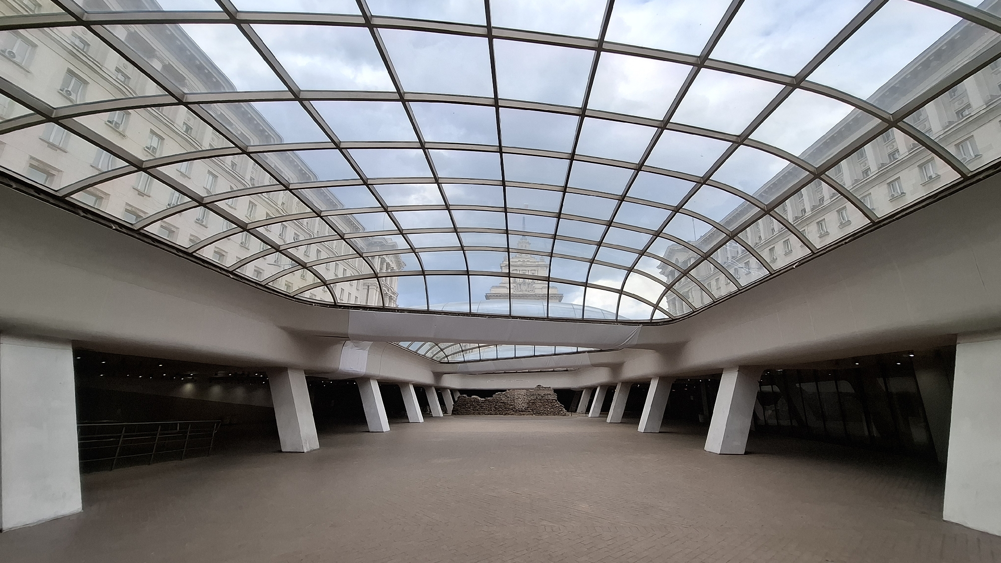 The image shows an interior space in Sofia, Bulgaria, featuring a modern architectural design with a curved glass ceiling supported by a grid of metal beams. The ceiling reflects the surrounding buildings and the sky above. Below the ceiling, there's a wide open area with a light brown, brick-patterned floor. The space is supported by large, white, trapezoidal columns. In the center of the open space is a small, fenced-off area containing exposed brick ruins. The background features dark, shadowed passageways that appear to lead to underground spaces. The image focuses on the architectural elements, with no people visible. The location is likely the Largo in Sofia, an area known for its mix of ancient ruins and modern structures.