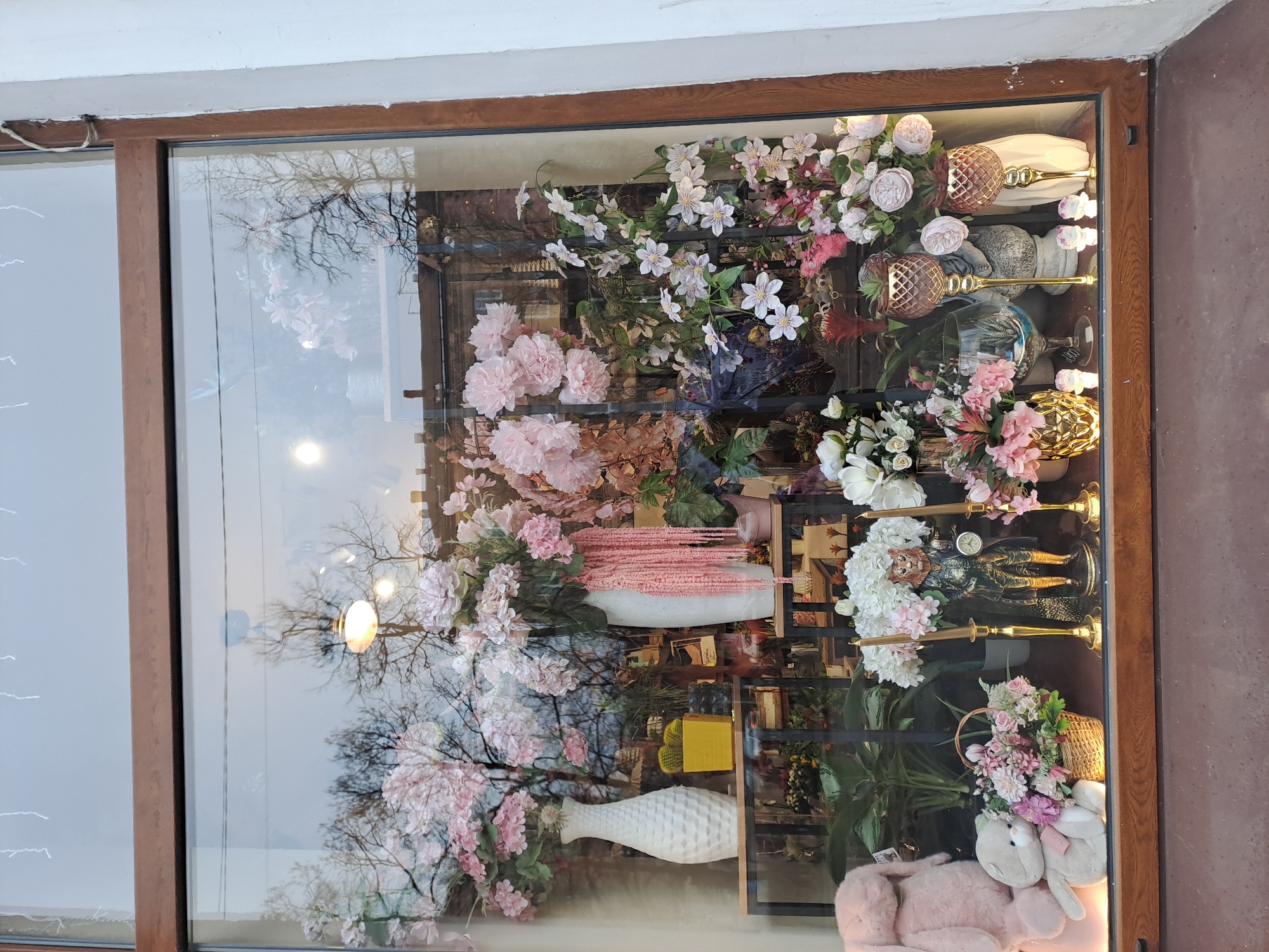 A window display in Мининская Поляна Кп, Russia, features a variety of floral arrangements, decorative vases, glassware, and figurines displayed on shelves. A stuffed bunny sits at the bottom of the display. A tree is reflected in the glass.
