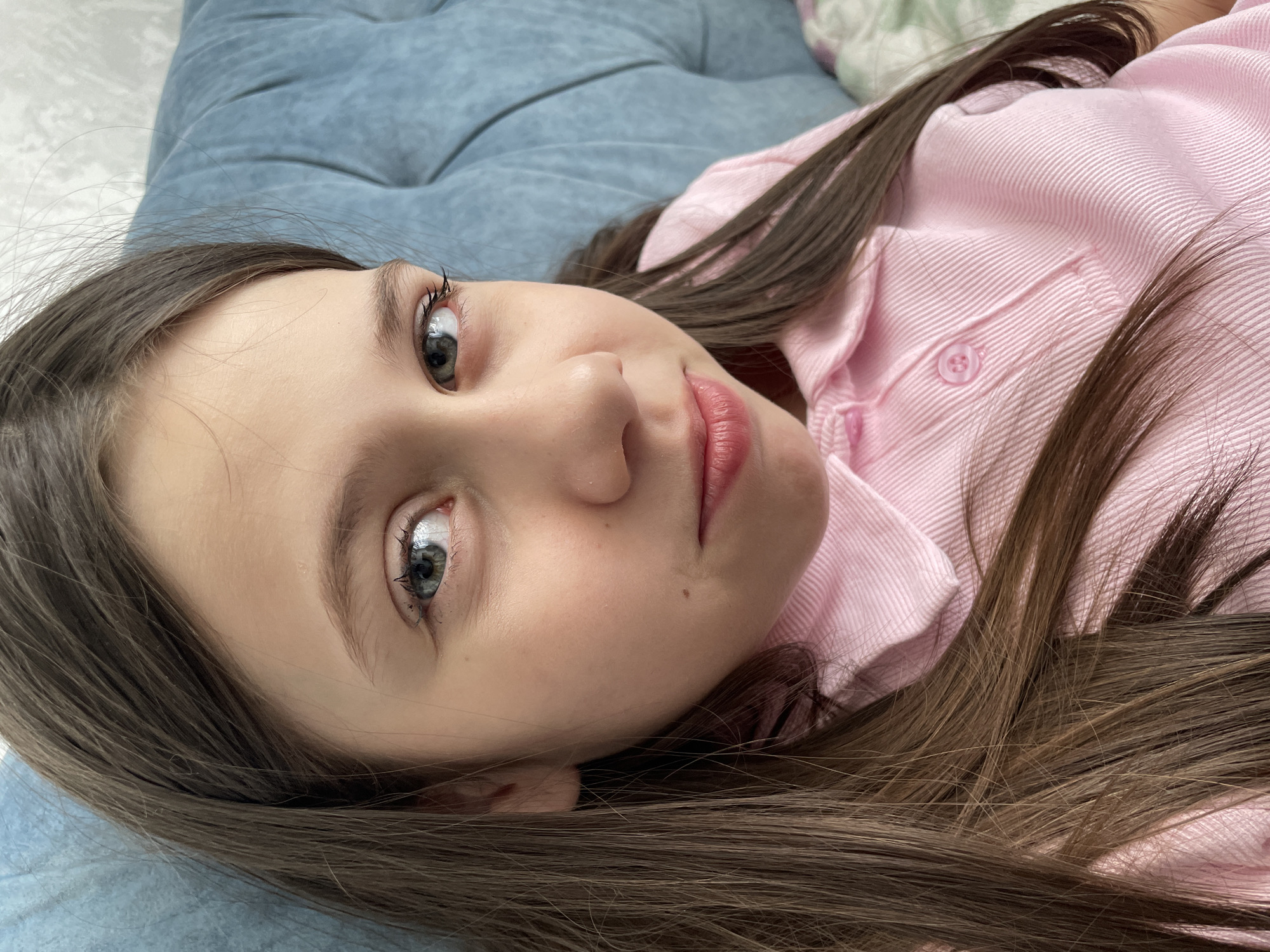 A young Caucasian girl with long brown hair and light eyes is lying on a blue surface, possibly a sofa or bed. She is wearing a light pink button-down shirt. The image is a close-up of her face and upper body, taken from a slightly angled, top-down perspective. The location in Izhevsk, Russia, is unknown from this image.