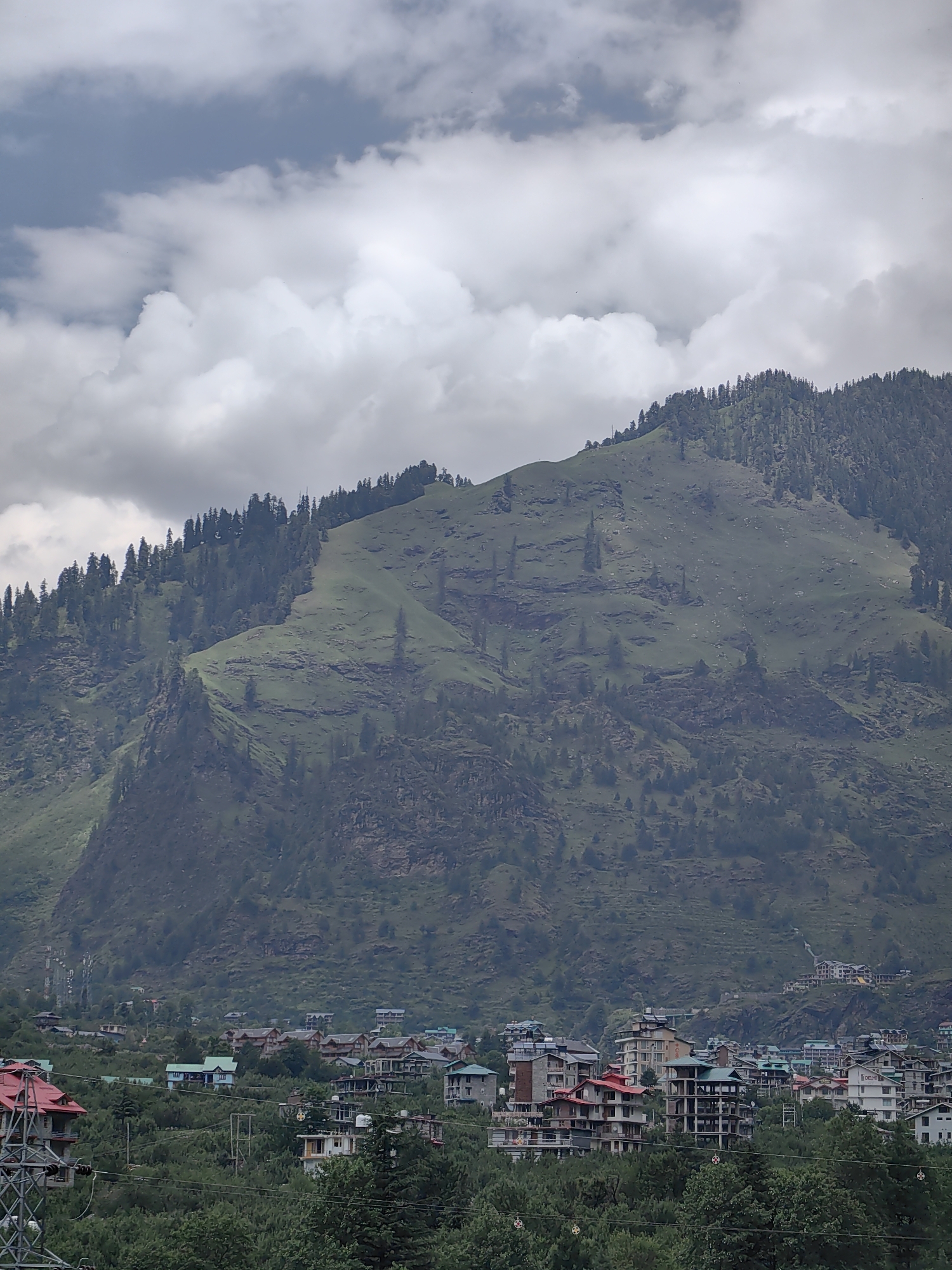 The image shows a view of the city of Kullu, India, nestled in a valley at the base of a large, green mountain. The mountain is covered in a dense forest of pine trees, with some rocky outcrops visible. The sky is cloudy, with patches of blue peeking through.
In the foreground, there are several buildings, mostly of a modest size and architectural style typical of residential areas in the region. Some of these buildings appear to be hotels or guesthouses, indicated by their multi-story structures and balconies. The architecture is characterized by pitched roofs, often in red or green, and light-colored facades.
The city extends up the lower slopes of the mountain, with buildings interspersed among trees. A winding road or path is visible on the mountain's flank. Electrical poles and wires are present, hinting at the infrastructure supporting the community. The overall scene depicts a tranquil, mountainous urban environment, suggesting a popular tourist destination due to its scenic beauty and proximity to nature. No people are visible in the image.