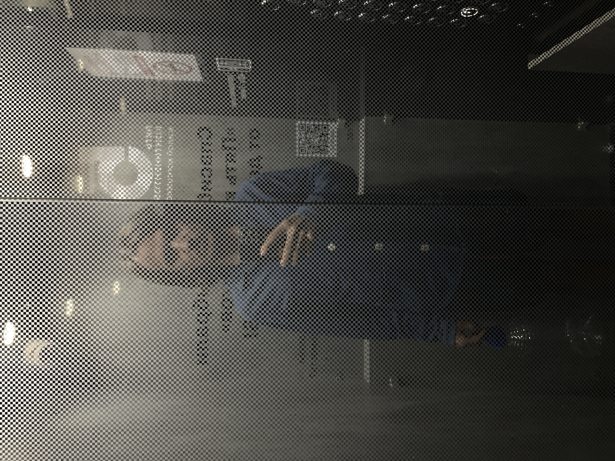 A full-body, front-facing mirror selfie of a person taken through a patterned glass wall. The person is wearing a blue collared shirt and is holding their right hand up, possibly taking the picture. The background is obscured by the patterned glass, but hints of an indoor environment with lighting and some signage are visible. The overall lighting is dim.