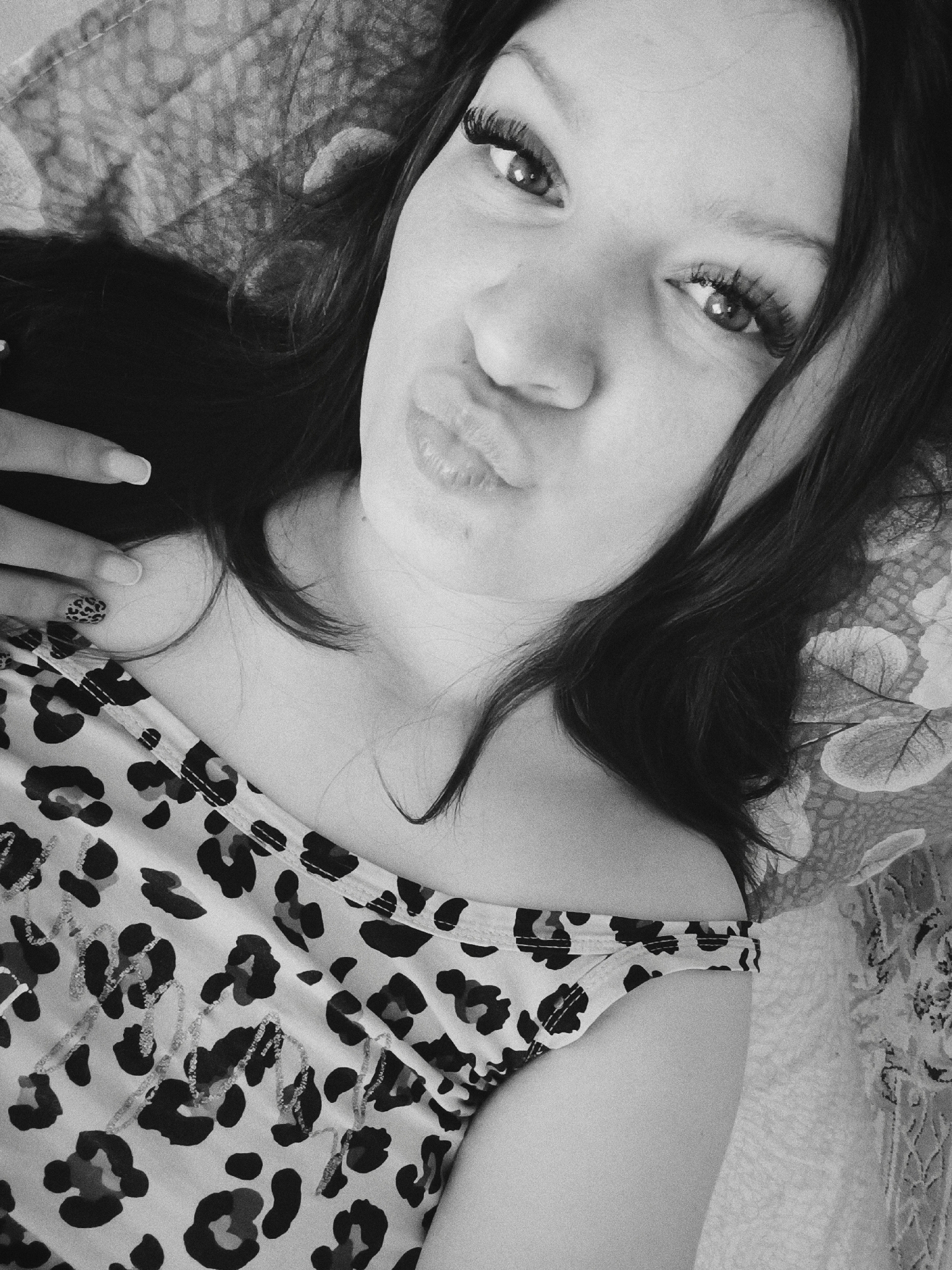 A selfie taken by a young woman with long, dark hair. She is lying down, looking directly at the camera with a slight pout. She is wearing a patterned top. Her hand is partially visible near her neck. The background appears to be a patterned bedspread or blanket. The image is in black and white. The location is not discernible from the image.