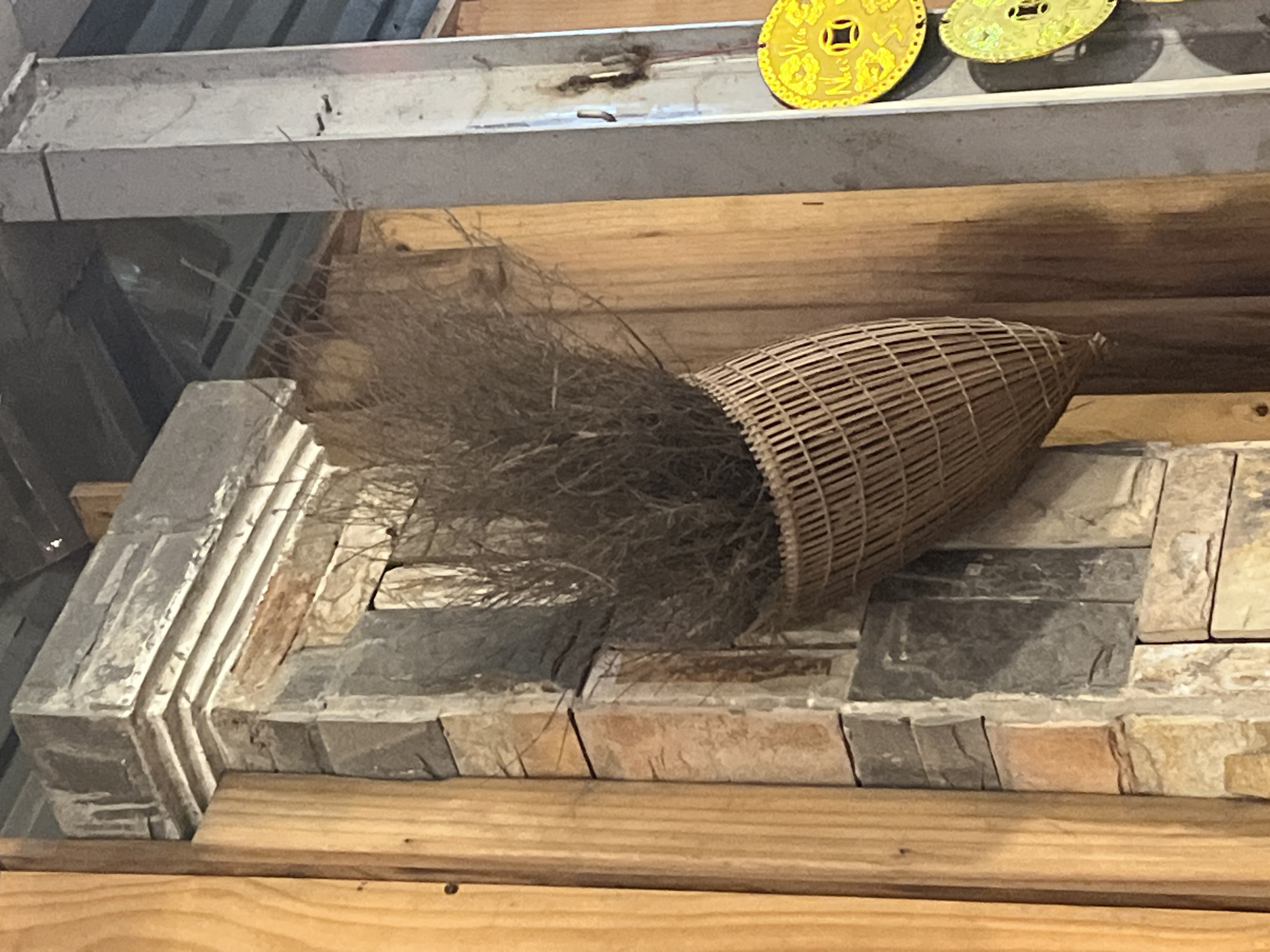 The image shows a close-up view of an exterior wall structure in Bac Nha Trang Ward, Vietnam. The wall features a pillar constructed from various stones, including rough-hewn, irregularly shaped grey and tan stones. A decorative, basket-like object woven from bamboo or similar material hangs from the pillar, filled with dry, brown reeds or grasses. To the right of the pillar, a section of a light grey, possibly metal, support beam is visible, with a few small nails or screws embedded in it. Two bright yellow circular ornaments, resembling coins with intricate designs, are attached to this beam. The background consists of wooden planks and what appears to be a corrugated metal roof or ceiling. The lighting suggests an indoor or covered outdoor setting. No people or animals are present in the frame.