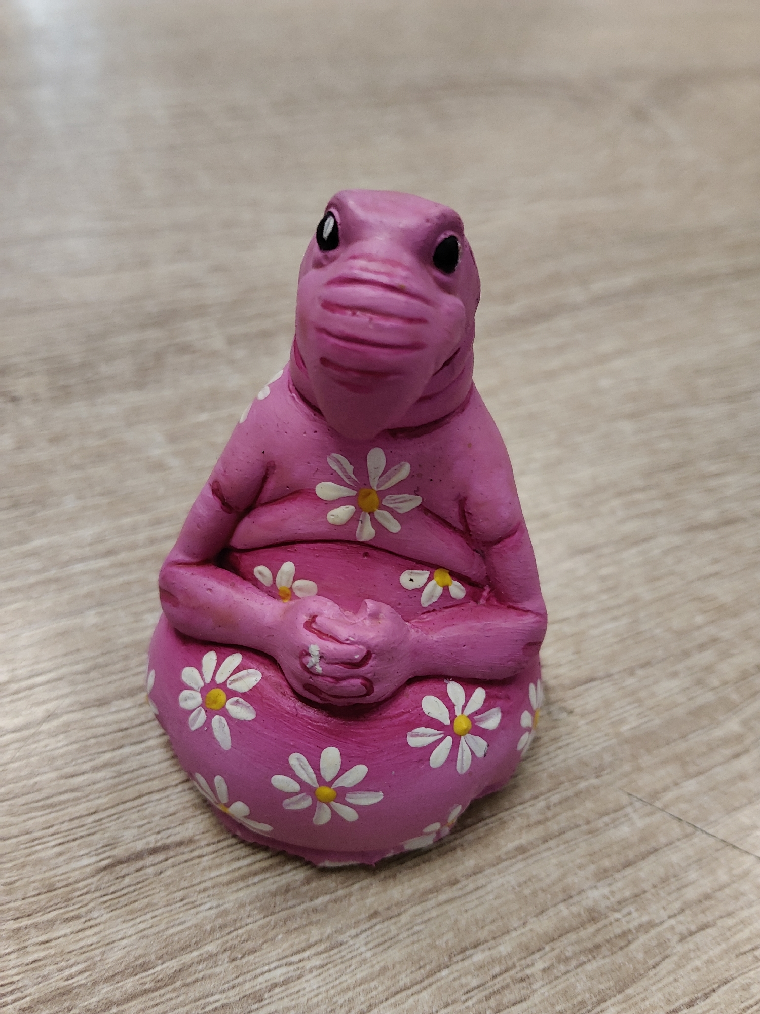 A pink, plump figurine sits on a light wood-grain surface. The figurine has a reptilian-like head with black eyes and a snout. Its body is decorated with white daisy-like flowers with yellow centers. The figurine's hands are clasped in its lap. The surface it rests on appears to be a table or floor. There is no indication of the city of Tver, Russia, in the image.