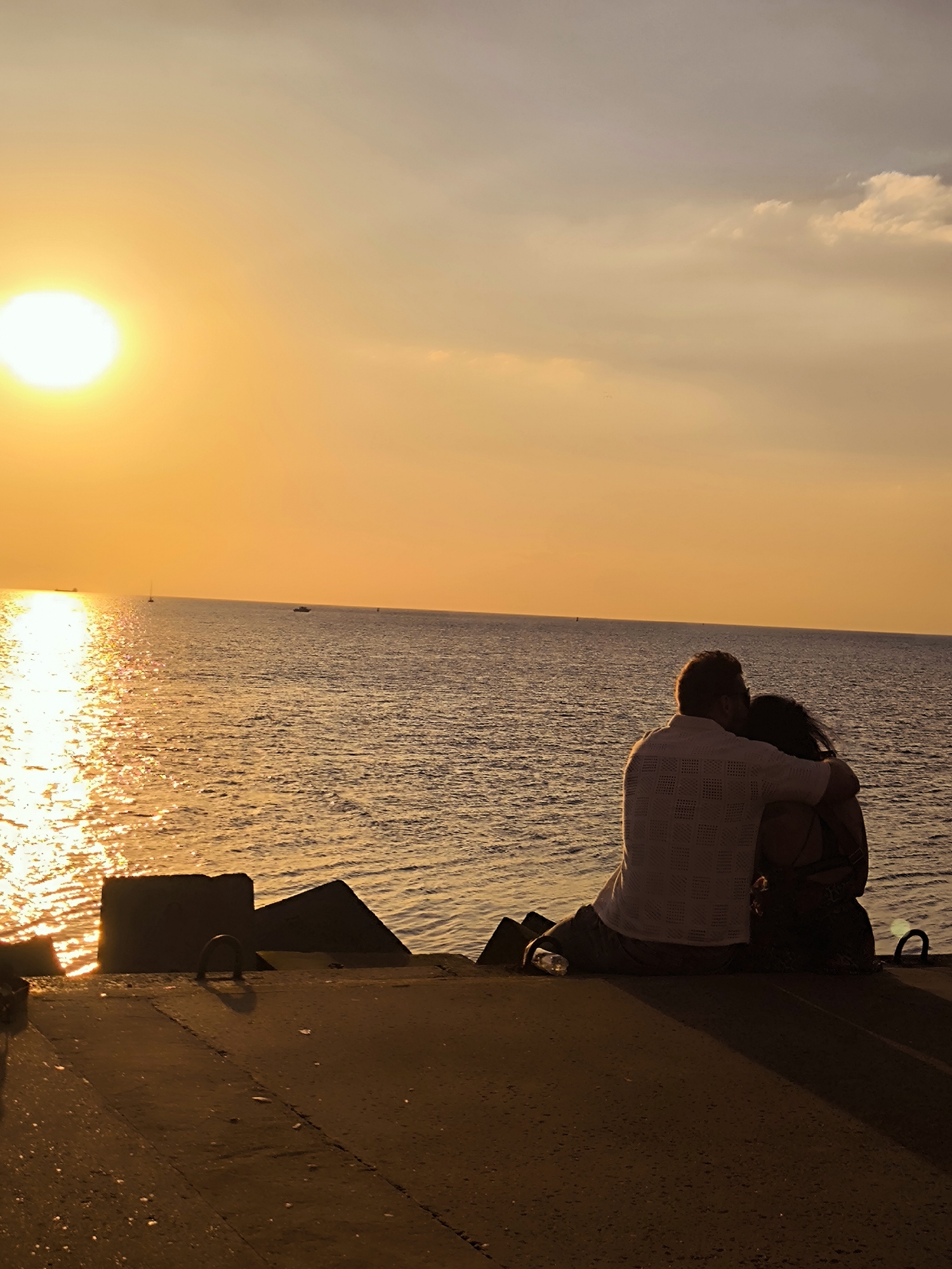 A couple embraces on a concrete structure overlooking a body of water at sunset. The sun, a bright orb, casts a golden glow across the sky and water, creating a shimmering reflection. The couple, silhouetted against the sunset, are seated with the man's arm around the woman. In the distance, a few boats are visible on the water. The foreground is a concrete surface with some angled concrete blocks. The scene appears to be a pier or breakwater. This image was taken in the city of Riga, Latvia, likely near Daugava River or the Baltic Sea coast.