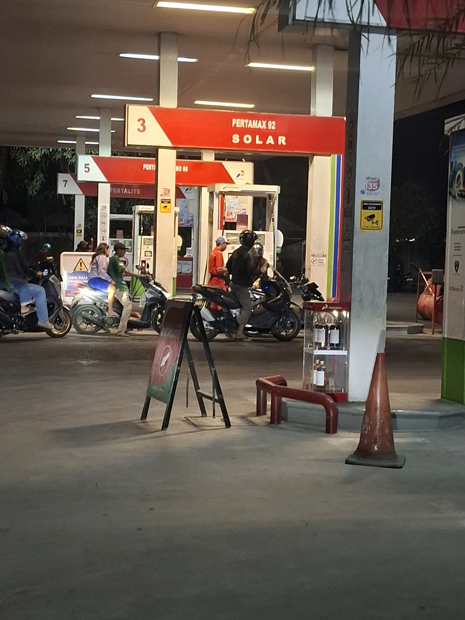 The image depicts a nighttime scene at a Pertamina gas station in Cibadak, Indonesia. The gas station features multiple fuel pumps under a covered canopy illuminated by fluorescent lights. Red and white signage is visible, with numbers "3", "5", and "7" indicating different service bays. One sign prominently displays "PERTAMAX 92 SOLAR". Several individuals are present, most of whom are on or near motorcycles. One man in an orange uniform appears to be a gas station attendant. Another man is operating a motorcycle while wearing a helmet. A couple is also seen riding together on a motorcycle. In the foreground, an A-frame sign with a red warning symbol is visible on the ground. Several motorcycles are parked or in use. To the right, a display cabinet contains bottles, and a traffic cone stands on the pavement. A CCTV sign is affixed to a pillar. The environment is paved, consistent with a gas station forecourt, and trees are visible in the background.