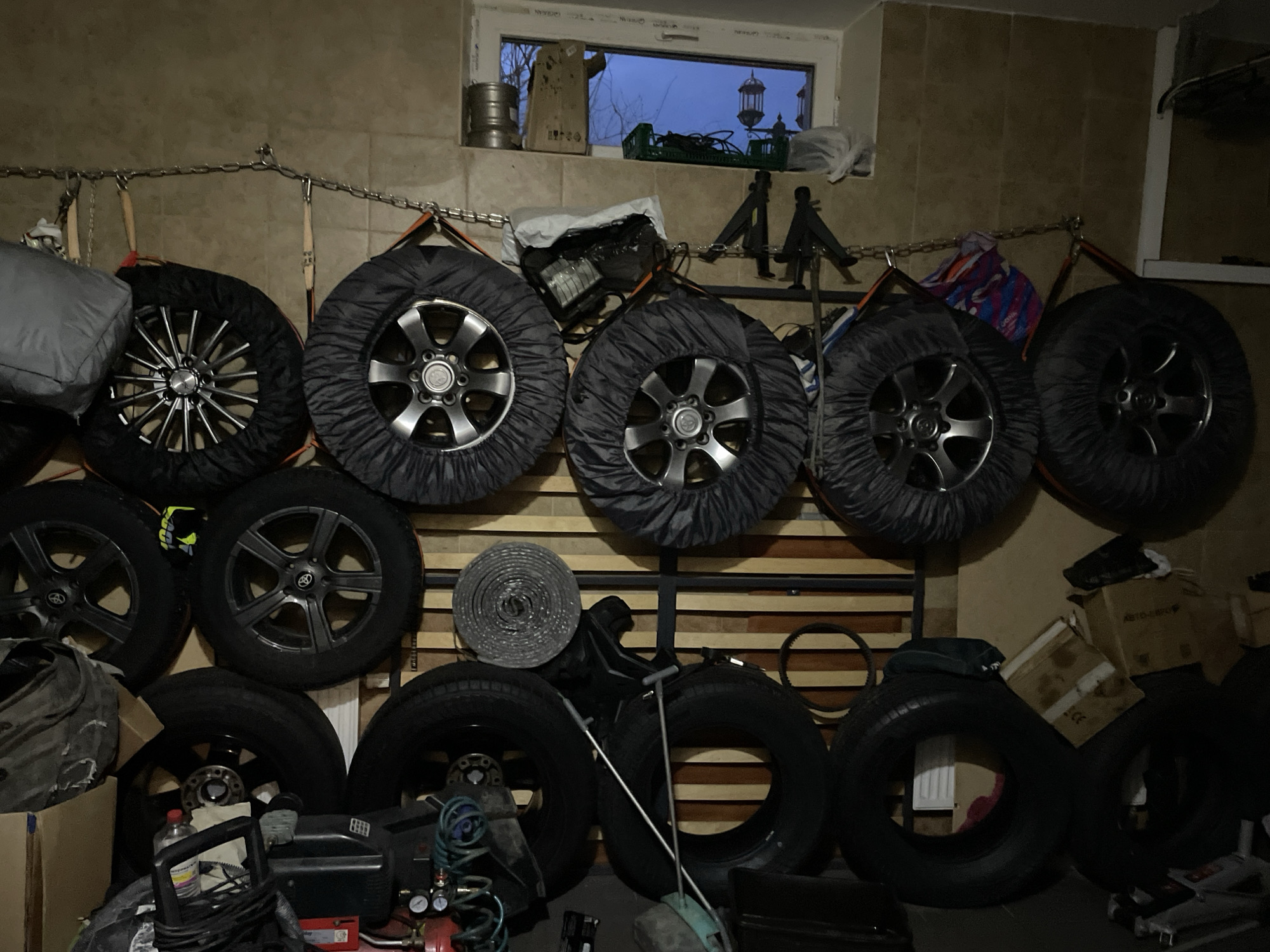 The image depicts an indoor space, possibly a garage or storage room, cluttered with various objects. A collection of car tires, some with covers, are mounted on a chain attached to the wall, suggesting a storage solution. Below the tires, on the floor, are more tires, tools, and equipment, including what appears to be an air compressor. There are also boxes and rolls of materials.
The location, Лесной Пейзаж Кп (шарапово), Russia, is provided, although there are no visual cues within the image to verify the precise setting. The environment appears to be dimly lit. No people are visible in the image.
