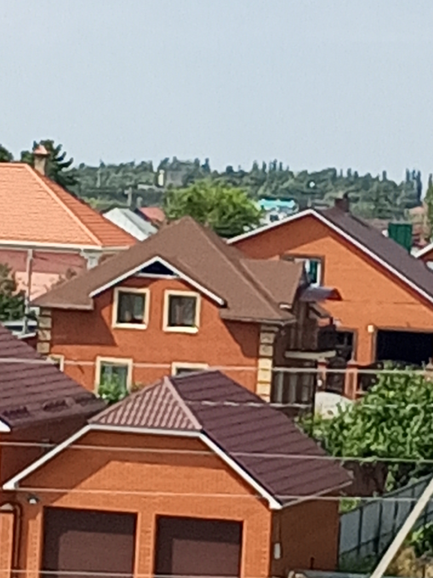 The image shows a residential area with several brick houses under a clear sky. The houses have varying roof designs, with some featuring brown tile roofs and others with a reddish-orange color. One house prominently displays two square windows on its upper floor, with a smaller rectangular window below. Another house in the foreground has a gabled roof and two large garage doors. Green trees and foliage are visible throughout the scene, adding to the suburban atmosphere. The overall impression is one of a quiet, sunny day in a neighborhood of single-family homes.