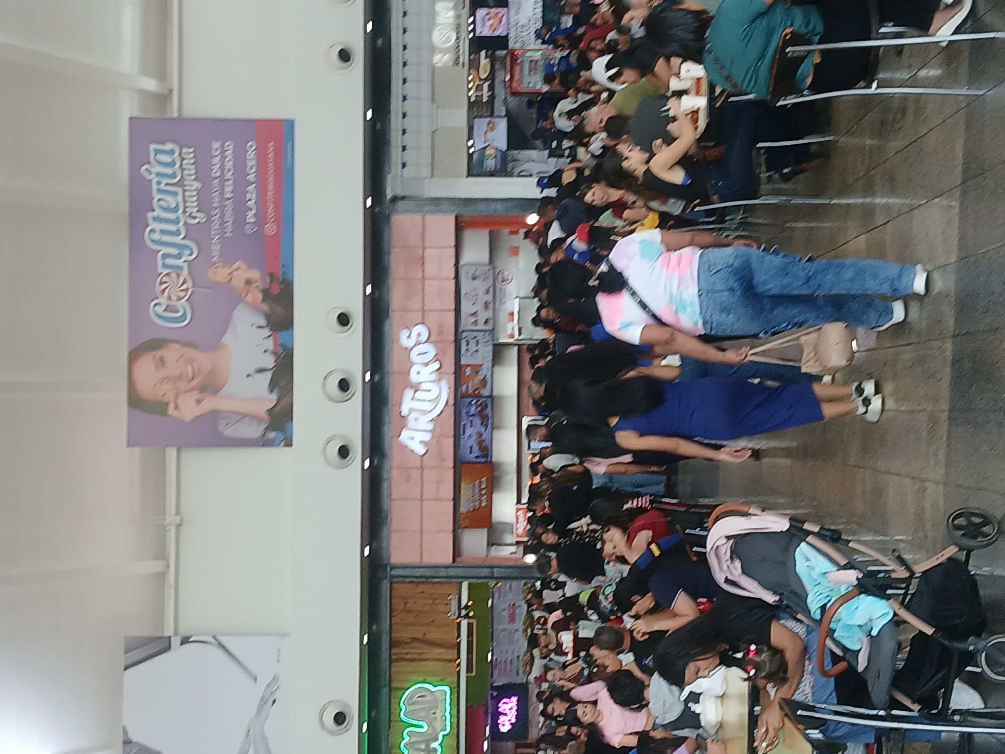 The image depicts a bustling indoor scene, likely a food court or a section of a shopping mall, filled with numerous people. The perspective is from an elevated vantage point, looking down at the crowd.
The primary subjects are a large number of individuals, including adults and children, populating the area. Many are standing in lines or congregating, while others are seated at tables in what appears to be dining areas. In the foreground, a person wearing a tie-dye shirt and blue jeans is standing, holding a bag. Near them, a baby stroller is visible, containing a light blue cloth and some items, suggesting the presence of a young child, though the child itself is not clearly visible. Further back, people are seen sitting at tables, some eating, and others engaging in conversation. The scene suggests a busy commercial environment, possibly during peak hours.
Notable details include the bright, artificial lighting typical of indoor public spaces, indicating it is likely daytime or early evening indoors. The atmosphere appears lively and crowded.
Several visible texts provide location cues. A prominent purple banner on the left side reads "Confitería Guayana" (Guayana Confectionery), followed by the slogan "MIENTRAS HAYA DULCE HABRÁ FELICIDAD" (While there is sweetness, there will be happiness). Below that, it states "PLAZA ACERO" (Steel Plaza) and a social media handle "@CONFITERIAGUAYANA". The mention of "Guayana" and "Plaza Acero" strongly links the setting to Ciudad Guayana, Venezuela, as indicated by the provided context. Another sign for a food establishment partially reads "ARTUROS" (or "ARTUR"). Other signs for various vendors are visible in the background, though their text is largely unreadable due to distance and angle. The architecture features multiple shopfronts with various food options, suggesting a diverse culinary offering.