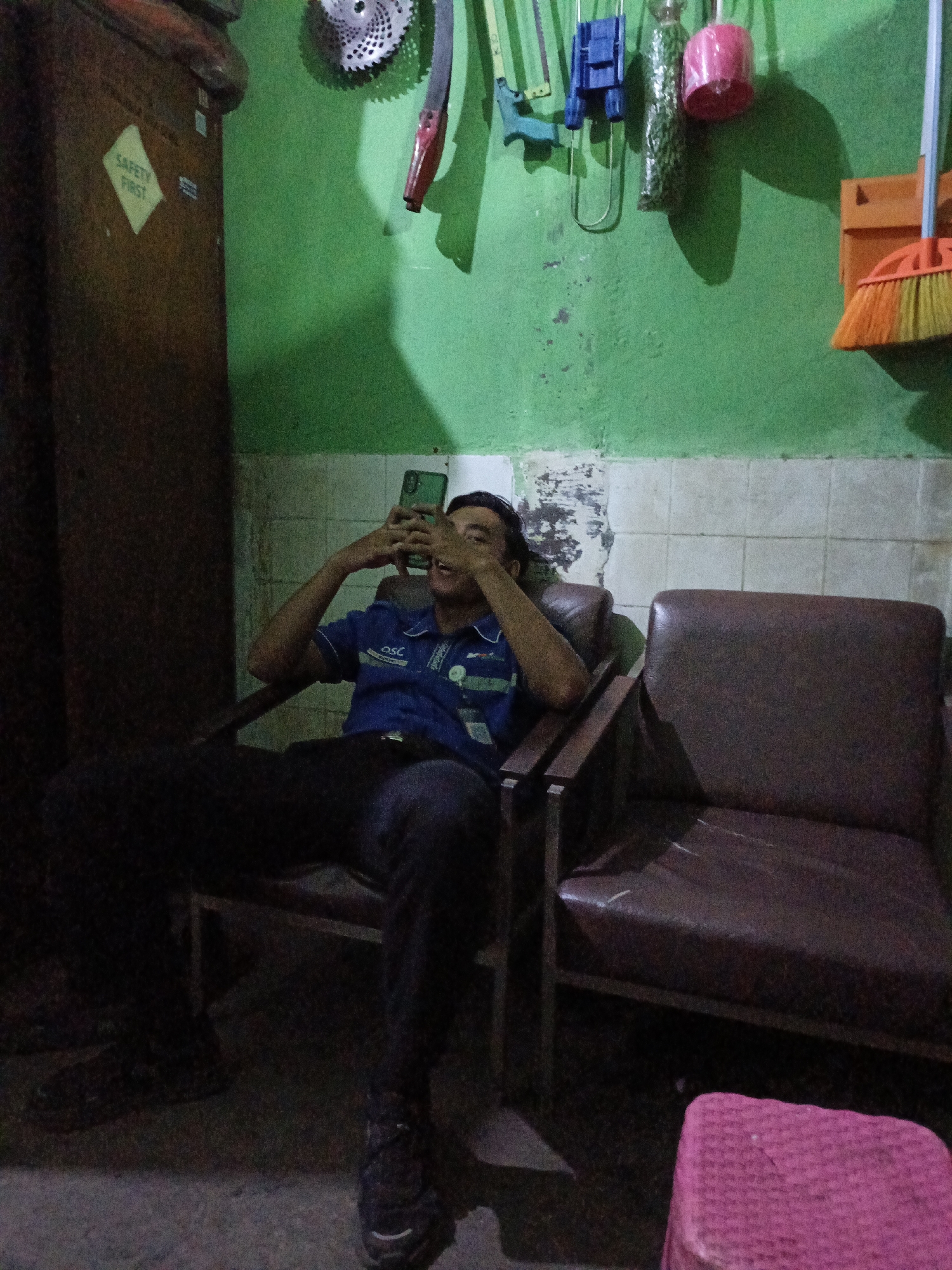 A young man sits in a chair, holding a phone. He is wearing a blue shirt and dark pants. The room has green walls and tiled floors. On the wall are various objects, including a broom and what appears to be a saw blade. A small pink stool is in the foreground. The image is taken in Kidul Dalem, Indonesia.