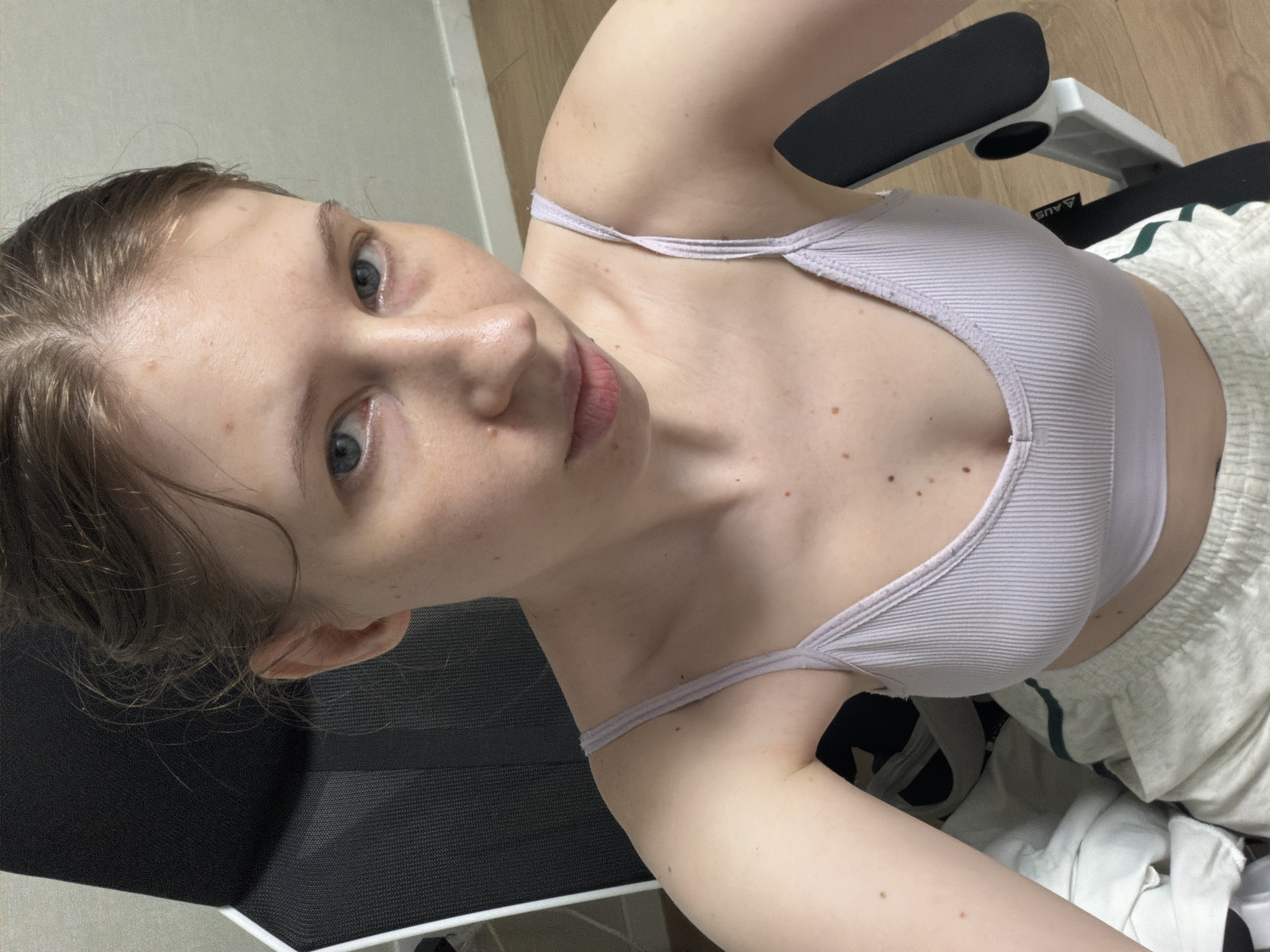 A young Caucasian woman with light skin and light brown hair tied back is captured from a high-angle, close-up perspective. She is wearing a light purple ribbed tank top and white shorts. Her eyes are a light blue-gray and she is looking directly at the camera. The background suggests an indoor setting, possibly a bedroom or living area, with wooden flooring and furniture including a black chair with white accents. The lighting appears to be natural, coming from an unseen source, and the time of day is indeterminate. No specific activity or event is clearly depicted, suggesting a casual, perhaps posed, photograph. There is no visible text in the image. The mood is neutral.