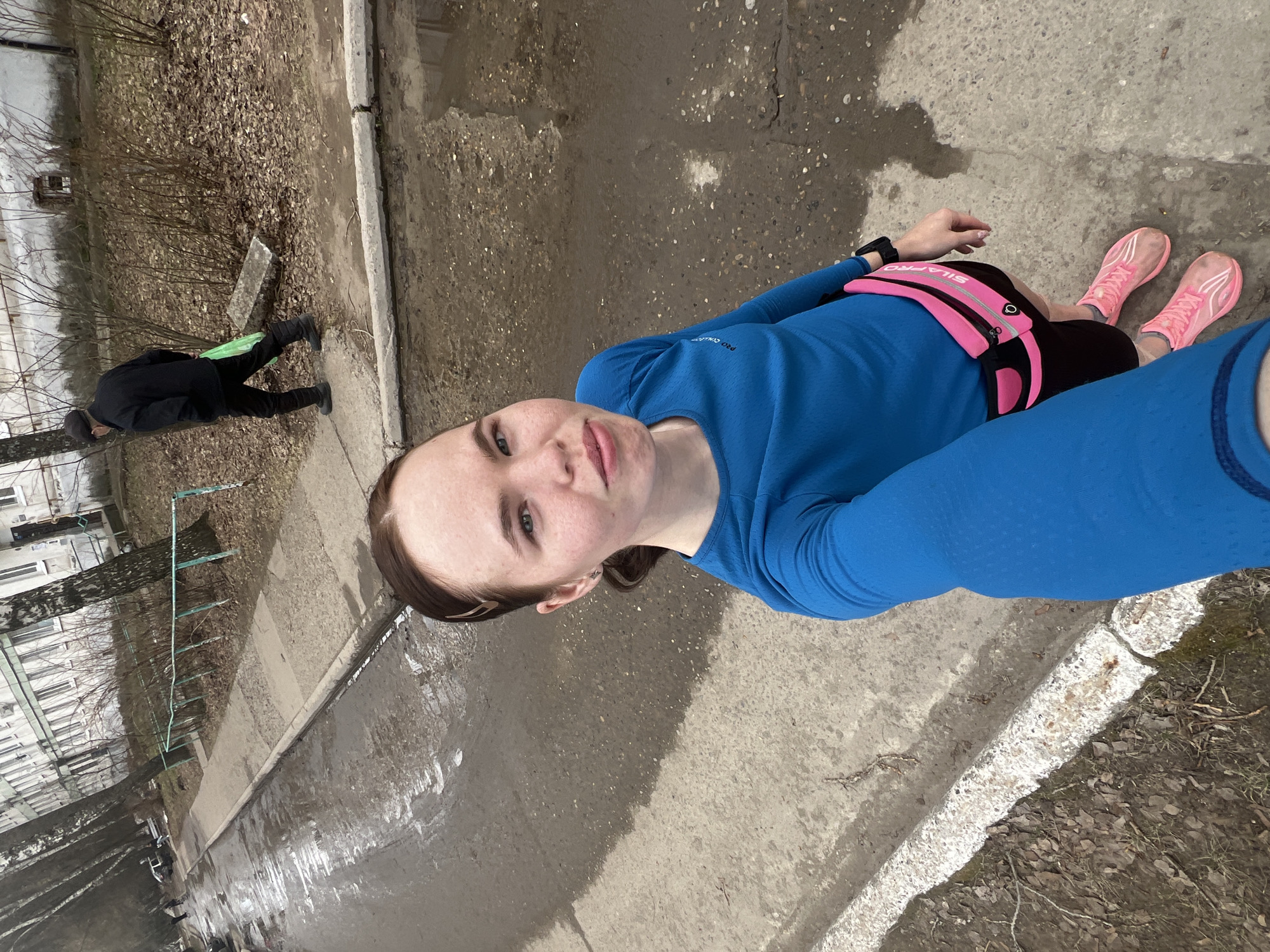 A person takes a selfie on what appears to be a street or sidewalk. The person is wearing a blue long-sleeved shirt, black shorts, and pink running shoes. They have a pink fanny pack around their waist. In the background, a person in dark clothing walks away from the camera, carrying a green object. There is a building visible in the background, along with trees and bare branches. The scene is located outdoors on a day with overcast lighting. The surface is wet, and there are patches of dry leaves and debris along the edge of the pavement. Based on the visual cues and metadata, the location is Chaikovsky, Russia.