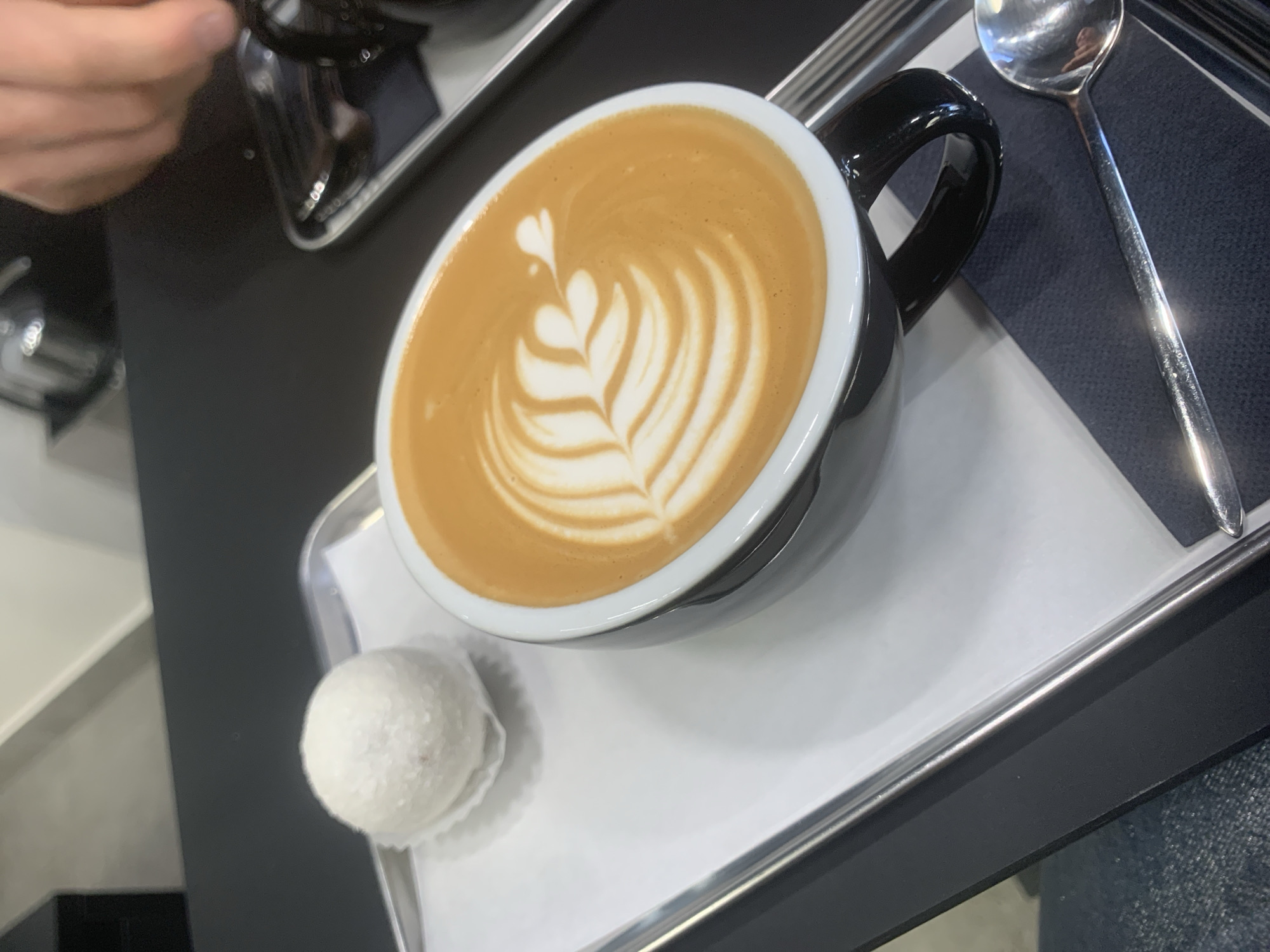 The image shows a cup of coffee with latte art on a silver tray. The cup is black and white, with a handle, and contains a light brown liquid topped with a white leaf pattern. To the left of the cup is a white, spherical confection. To the right of the cup, on the tray, is a silver spoon. The tray is lined with a white paper. The image appears to be taken from above at a slightly tilted angle. The background shows a glimpse of a dark table and what looks like a person's hand. The image does not include enough data to make definitive statements about location of the city of Izhevsk, Russia.