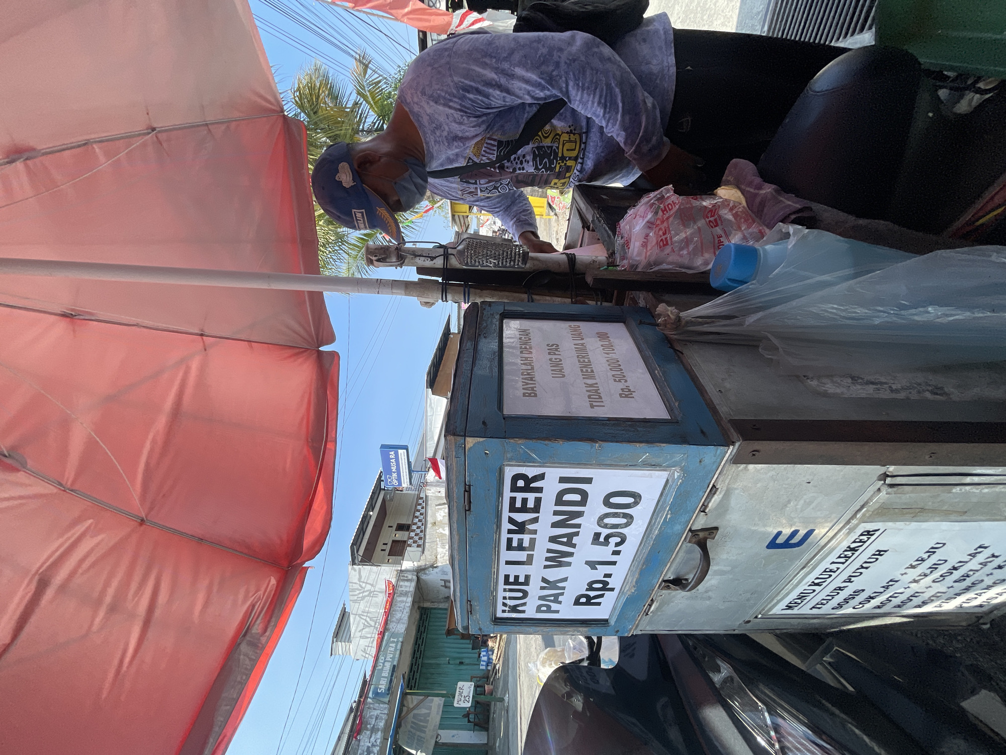 In Balonggabus, Indonesia, a vendor stands by a cart selling "Kue Leker Pak Wandi." The cart is a blue wooden box with a white sign displaying the name and price, Rp. 1.500. Another sign on the cart indicates payment instructions. The vendor, wearing a blue cap and a patterned long-sleeved shirt, is masked and appears to be serving a customer, though the customer is not visible in the frame. The scene is set outdoors under a large red umbrella. In the background, buildings and a street are visible, suggesting an urban setting. The overall atmosphere is that of a typical street food vendor.